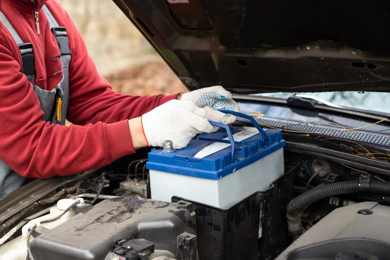 A person in a red sweater and gray coveralls wearing work gloves, lifting a car battery from an open vehicle hood.