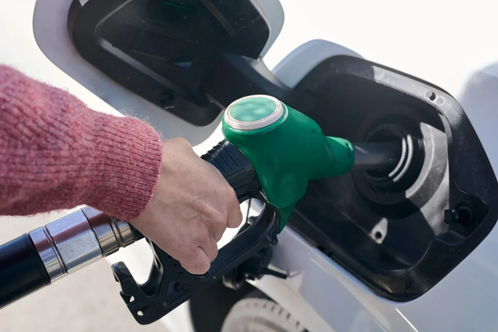 A hand in a pink sweater holds a green fuel nozzle while refueling a white vehicle.