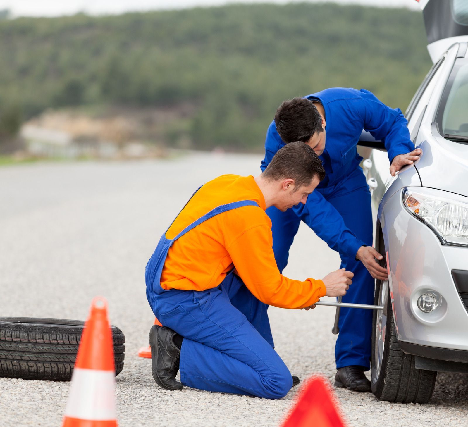 Two mechanics in blue uniforms, one wearing an orange shirt, change a car tire on the side of a road near orange cones.