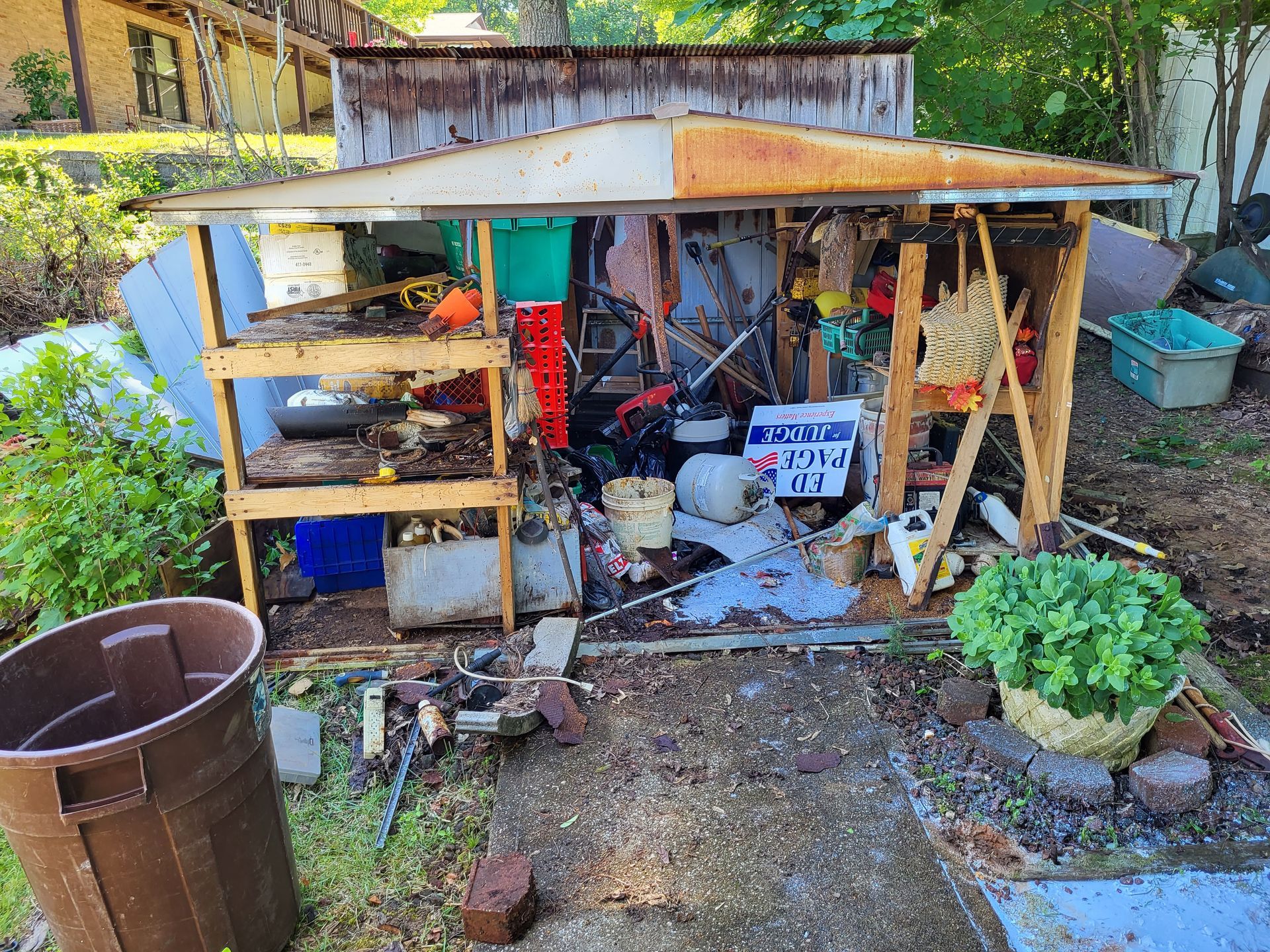 A shed filled with lots of junk and a trash can in front of it.