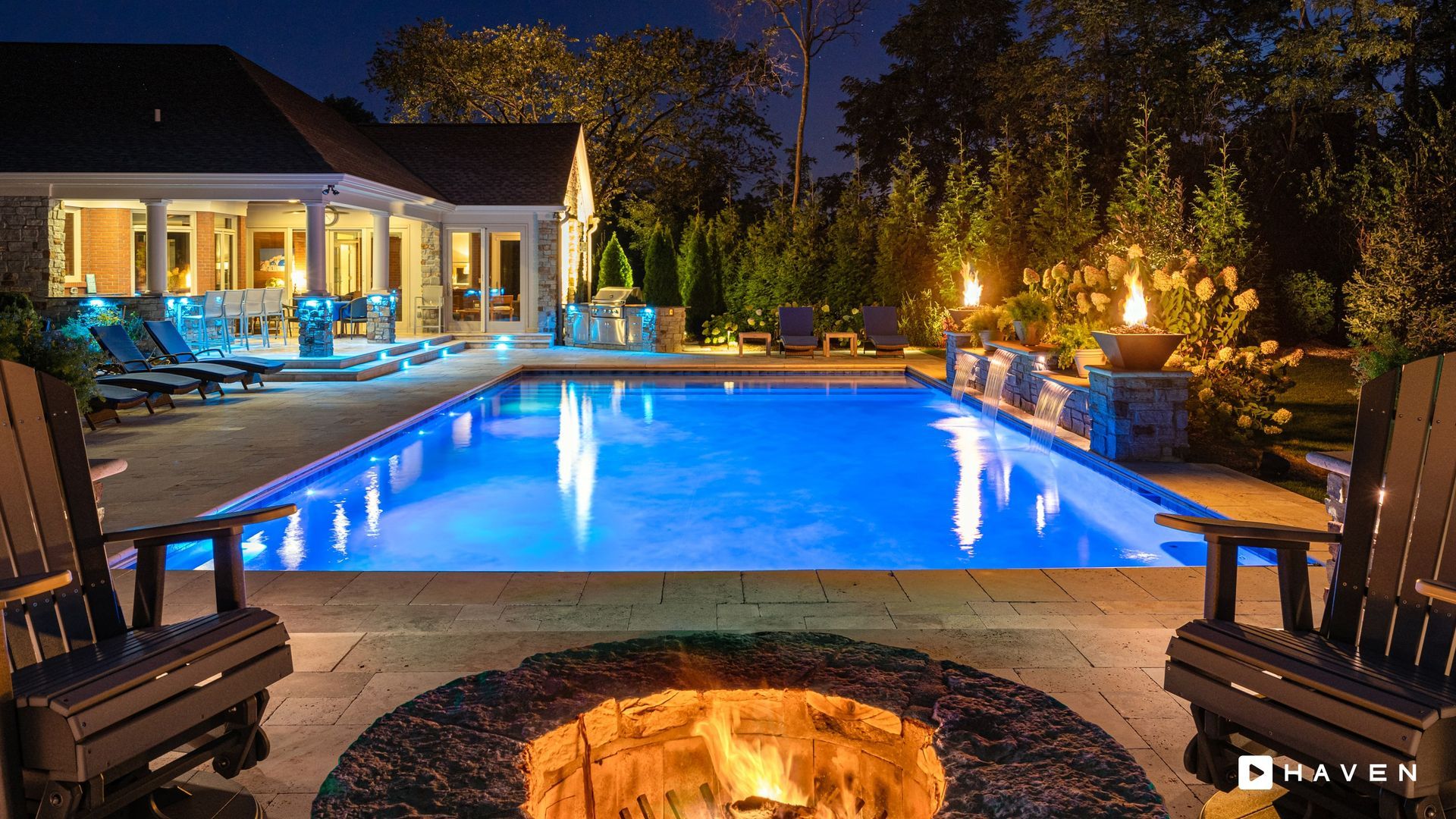 Backyard swimming pool at night with illuminated water, lounge chairs, outdoor grill area, and landscaped trees with accent lighting.