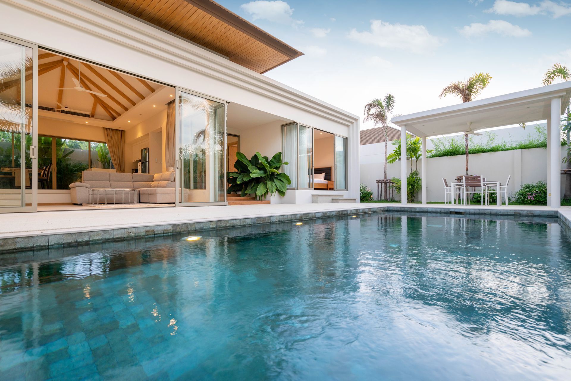 A turquoise pool in front of a modern white villa with a patio, outdoor dining area, and large sliding glass doors.