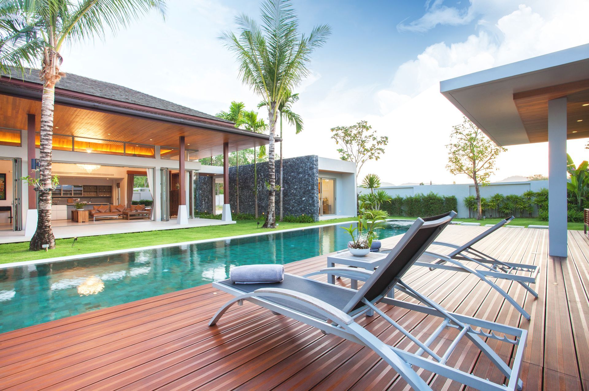 Modern luxury home exterior with a long swimming pool, wooden deck lounge chairs, palm trees, and covered outdoor living area.