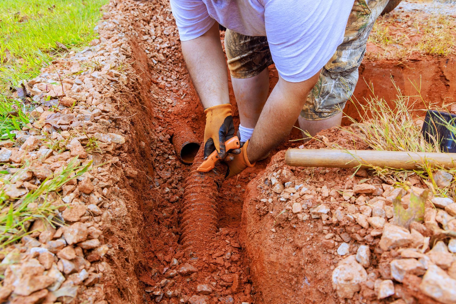 Person in gloves working on drainage pipes in a trench filled with reddish soil and gravel.