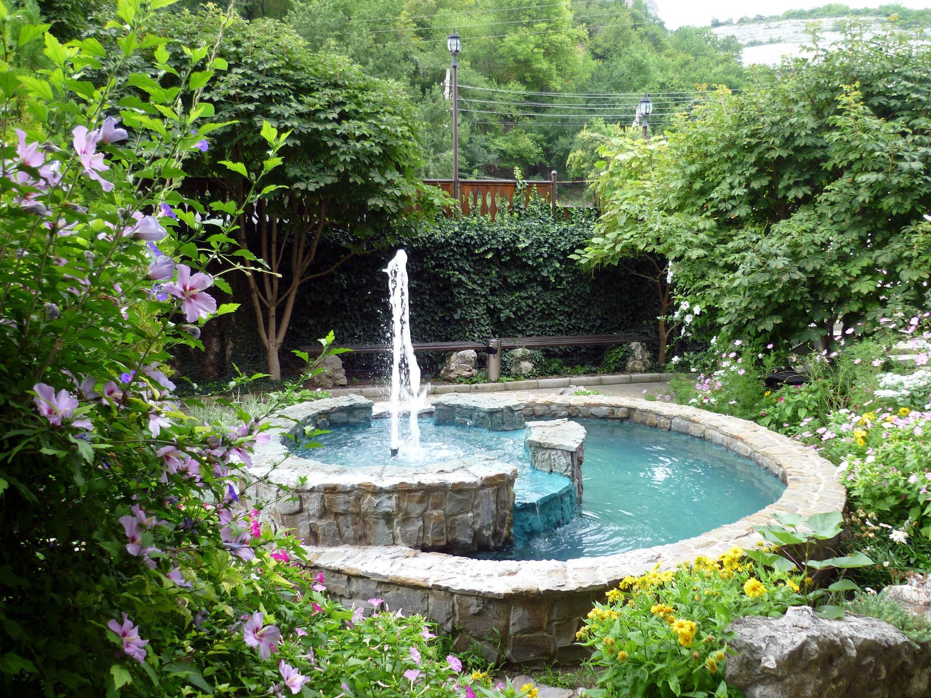 There is a fountain in the middle of the garden surrounded by flowers.