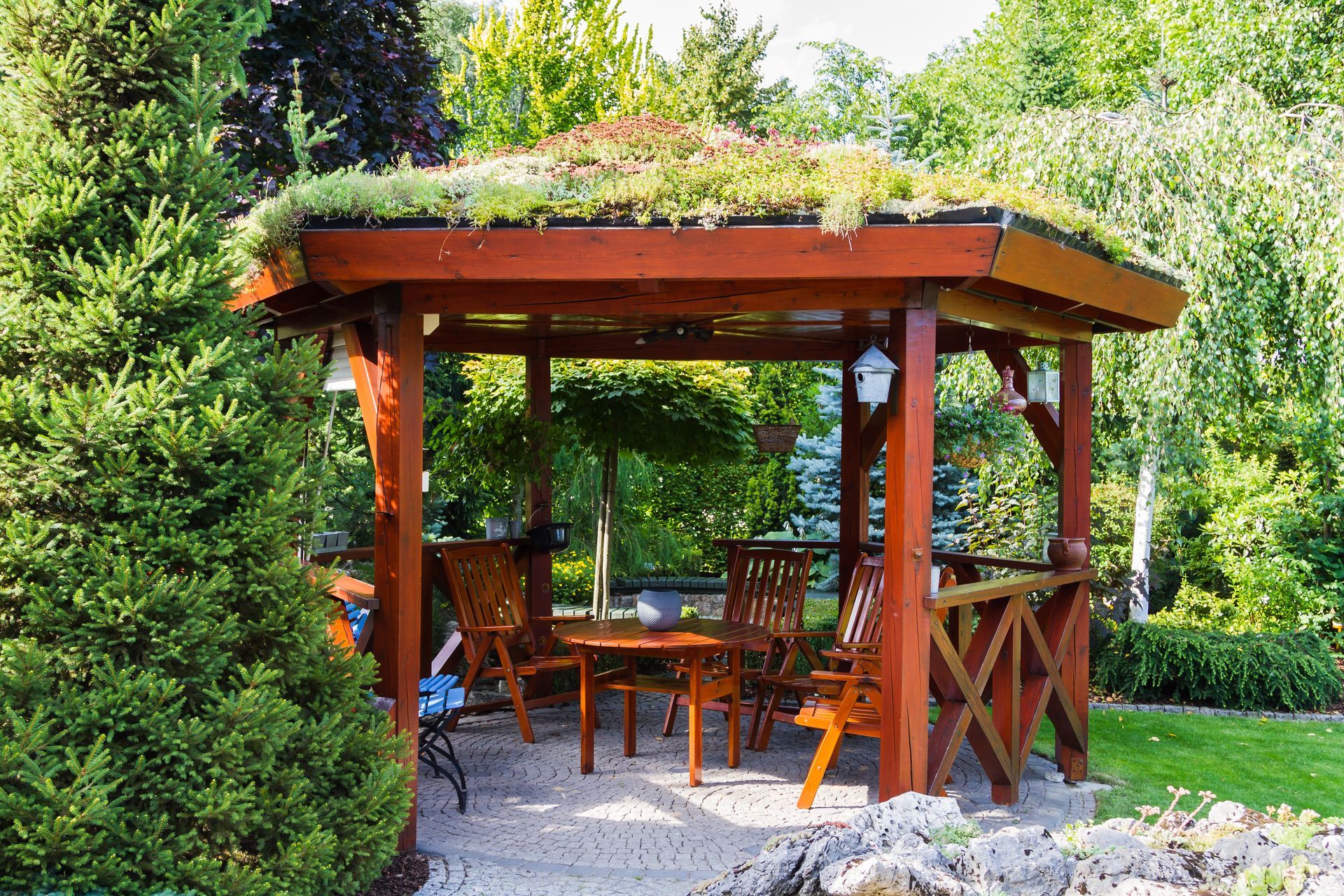 A wooden gazebo with a green roof and a table and chairs in a garden.