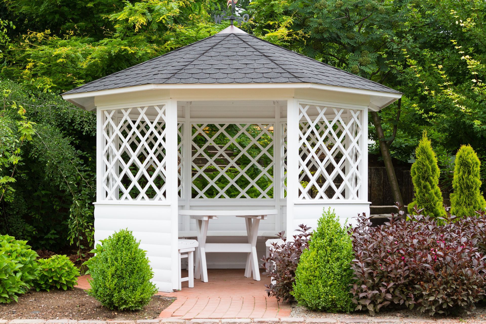 A white gazebo with a black roof is surrounded by trees and bushes.