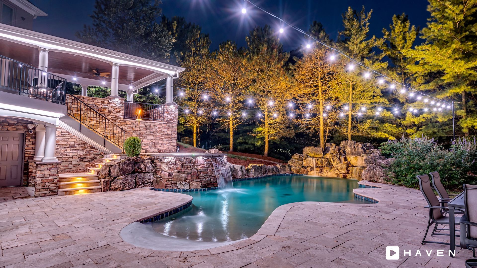A stone-tiled patio at night featuring an illuminated pool with a waterfall, stone walls, and string lights overhead.