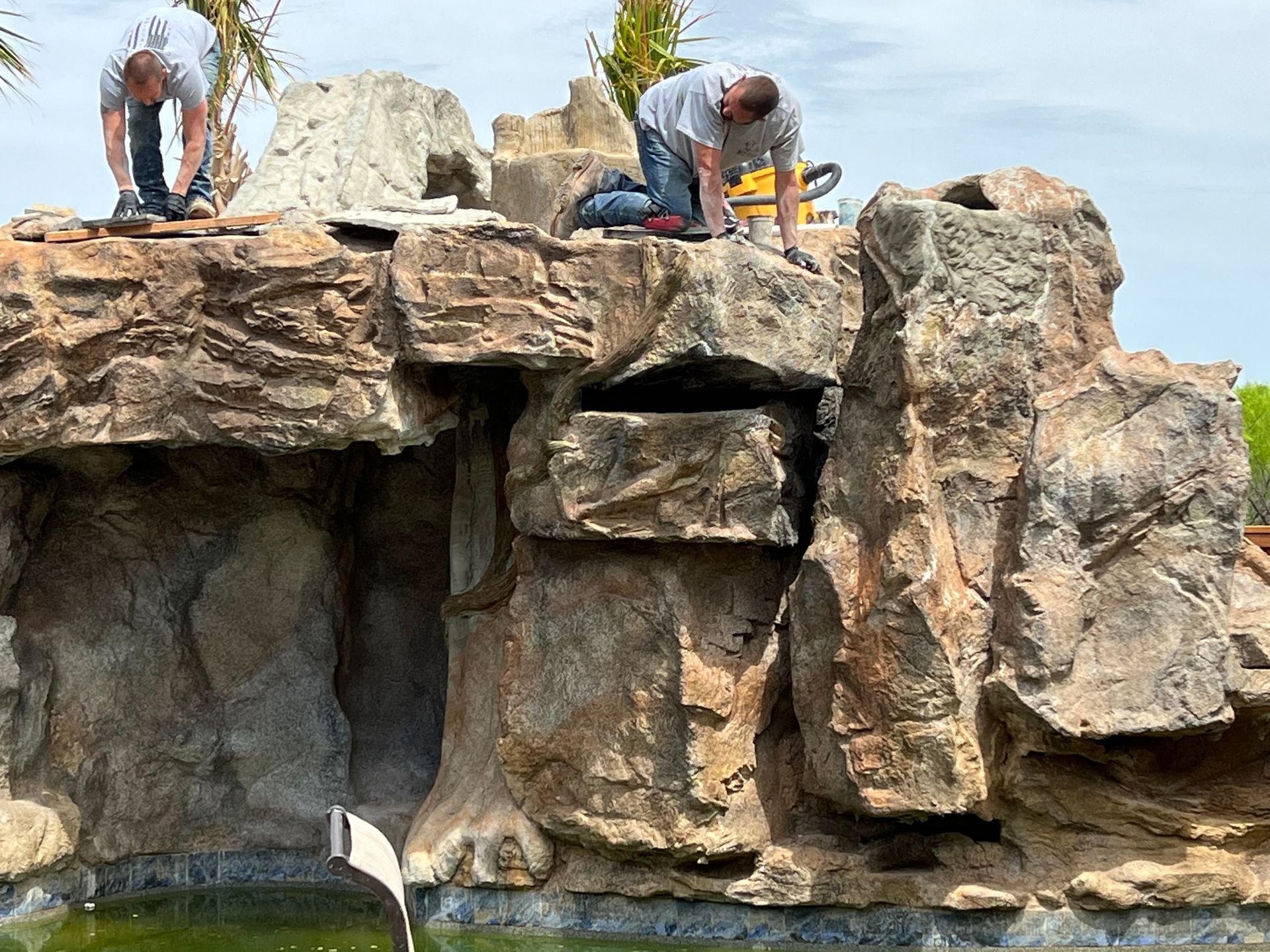 Two men are working on a large rock formation next to a pool.