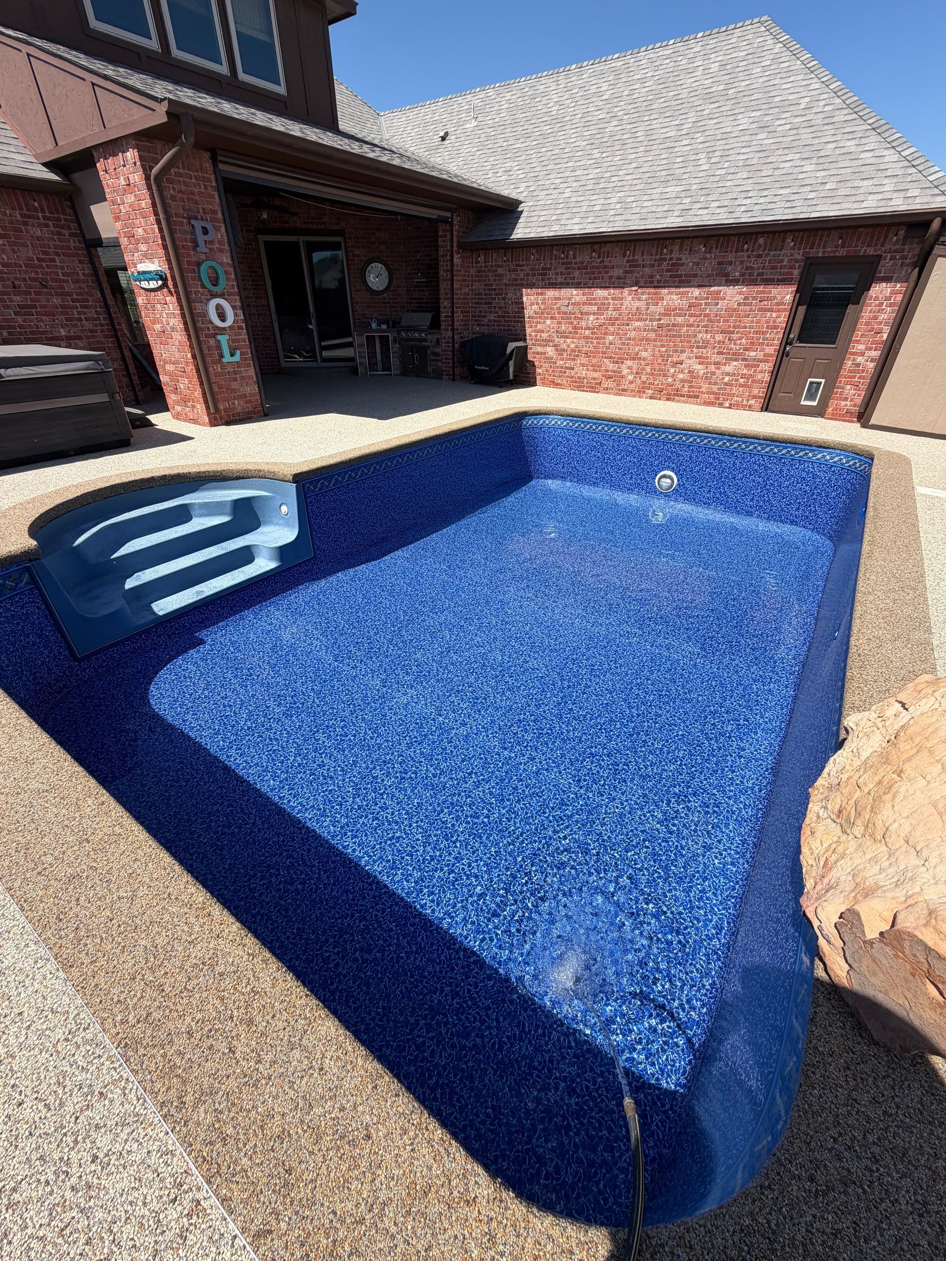 An empty, rectangular swimming pool with deep blue mosaic tiles and built-in steps next to a brick house patio.