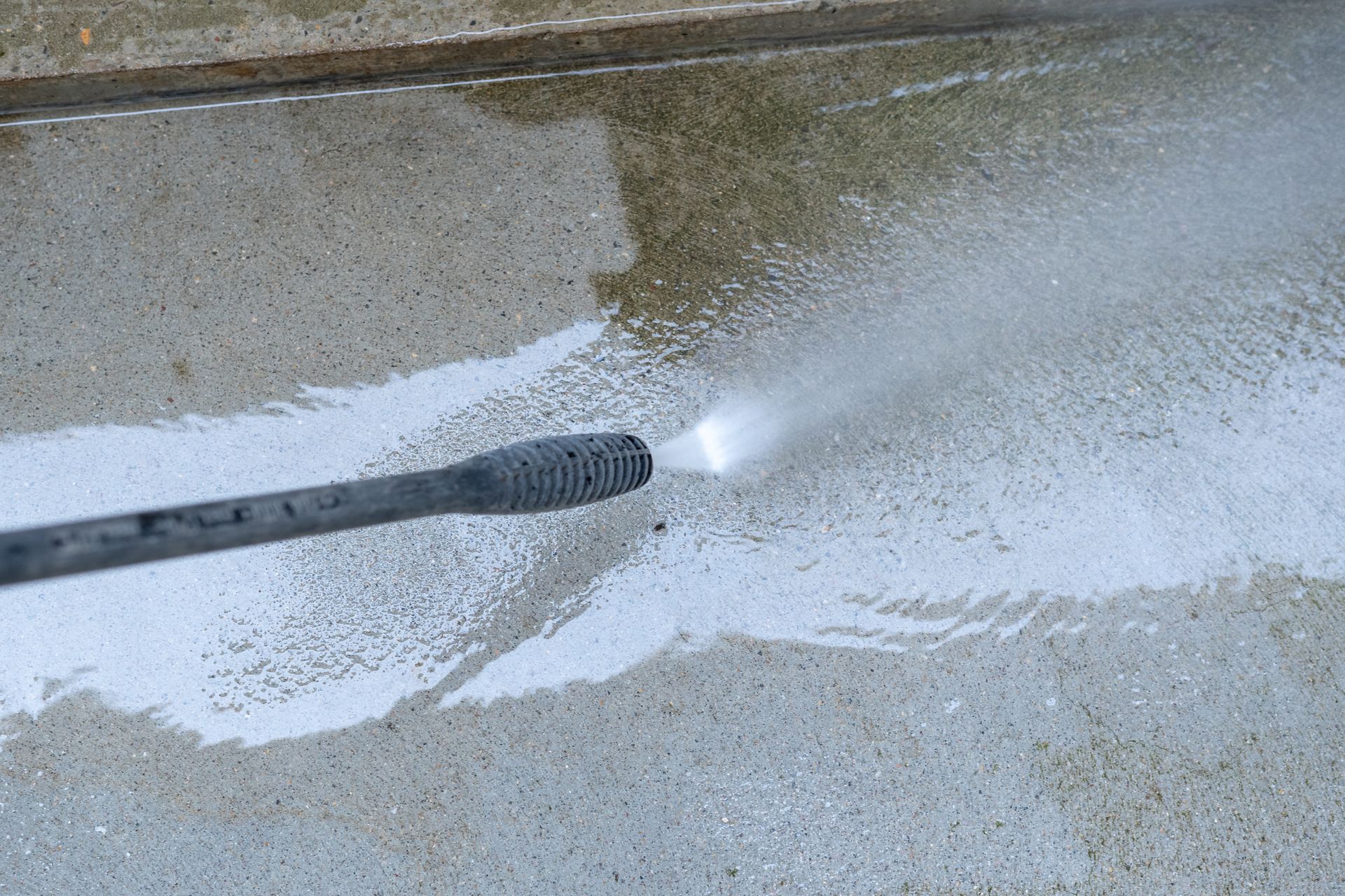 Elite Outdoor Living Solutions is pressure washing a dirty concrete driveway surface with a hose in Moore, OK.