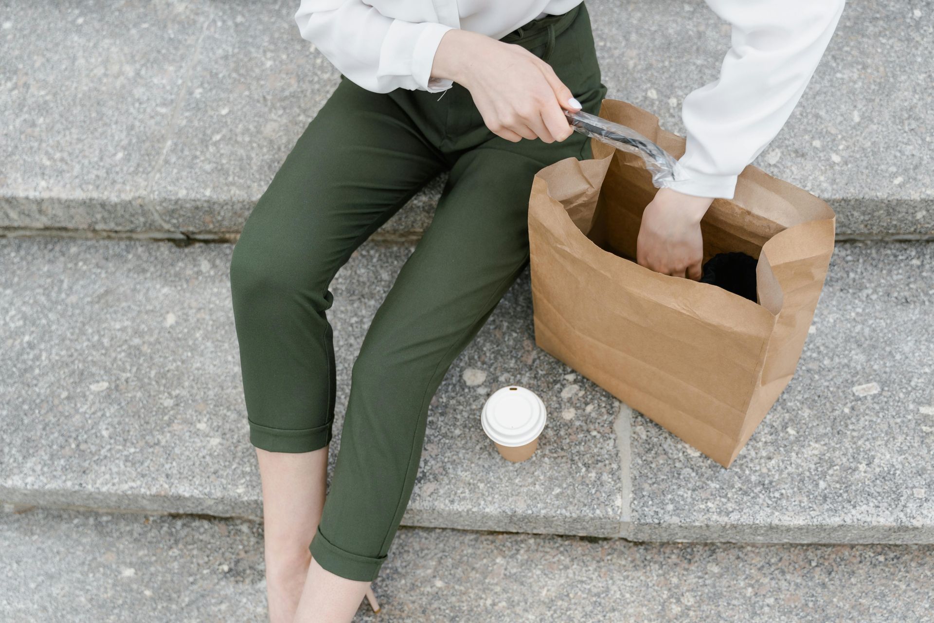 A woman is sitting on a set of stairs with a paper bag and a cup of coffee.