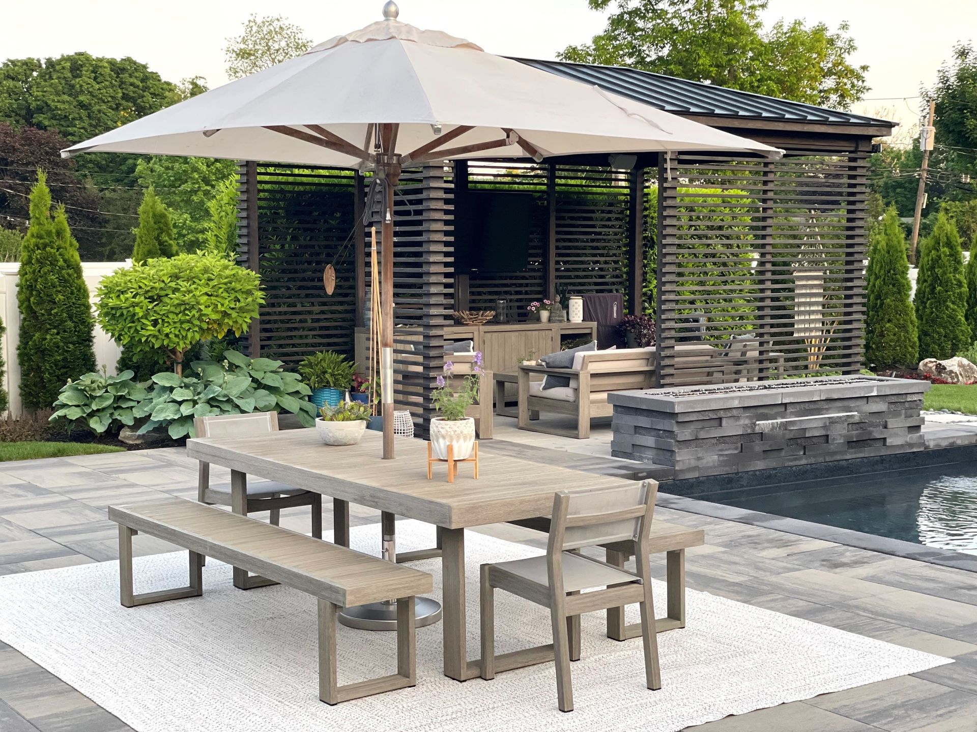 A patio with a table and benches under an umbrella next to a pool.