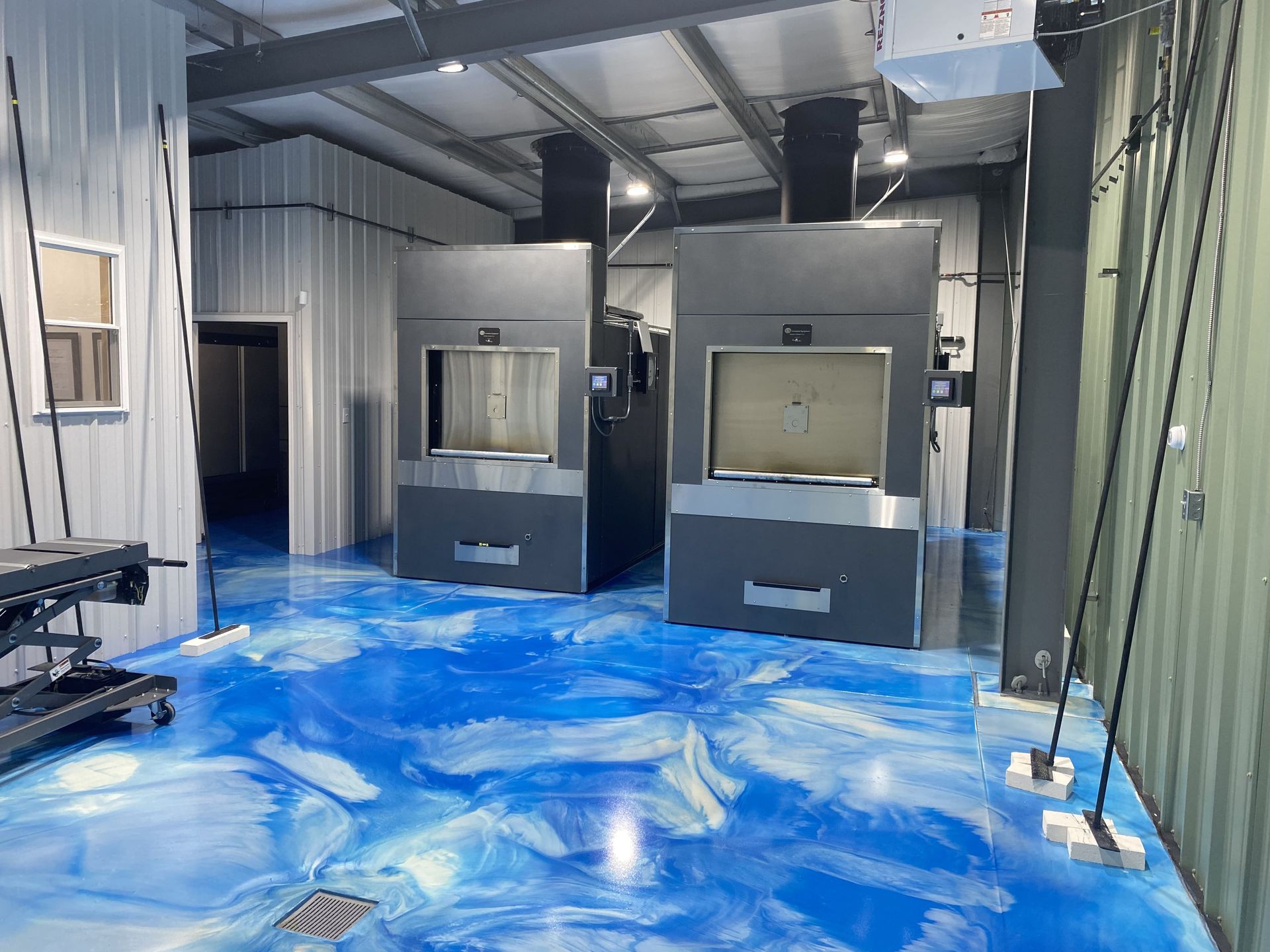 Two industrial incinerators in a room with blue epoxy flooring, metal beams, and a small window.