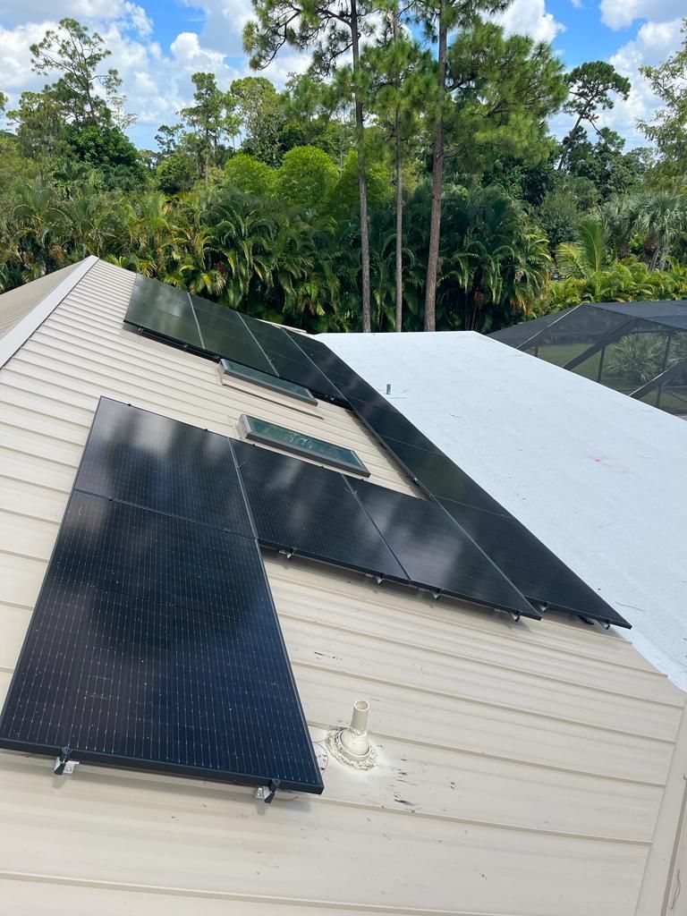 Rooftop with multiple black solar panels on a light-colored house, surrounded by trees.