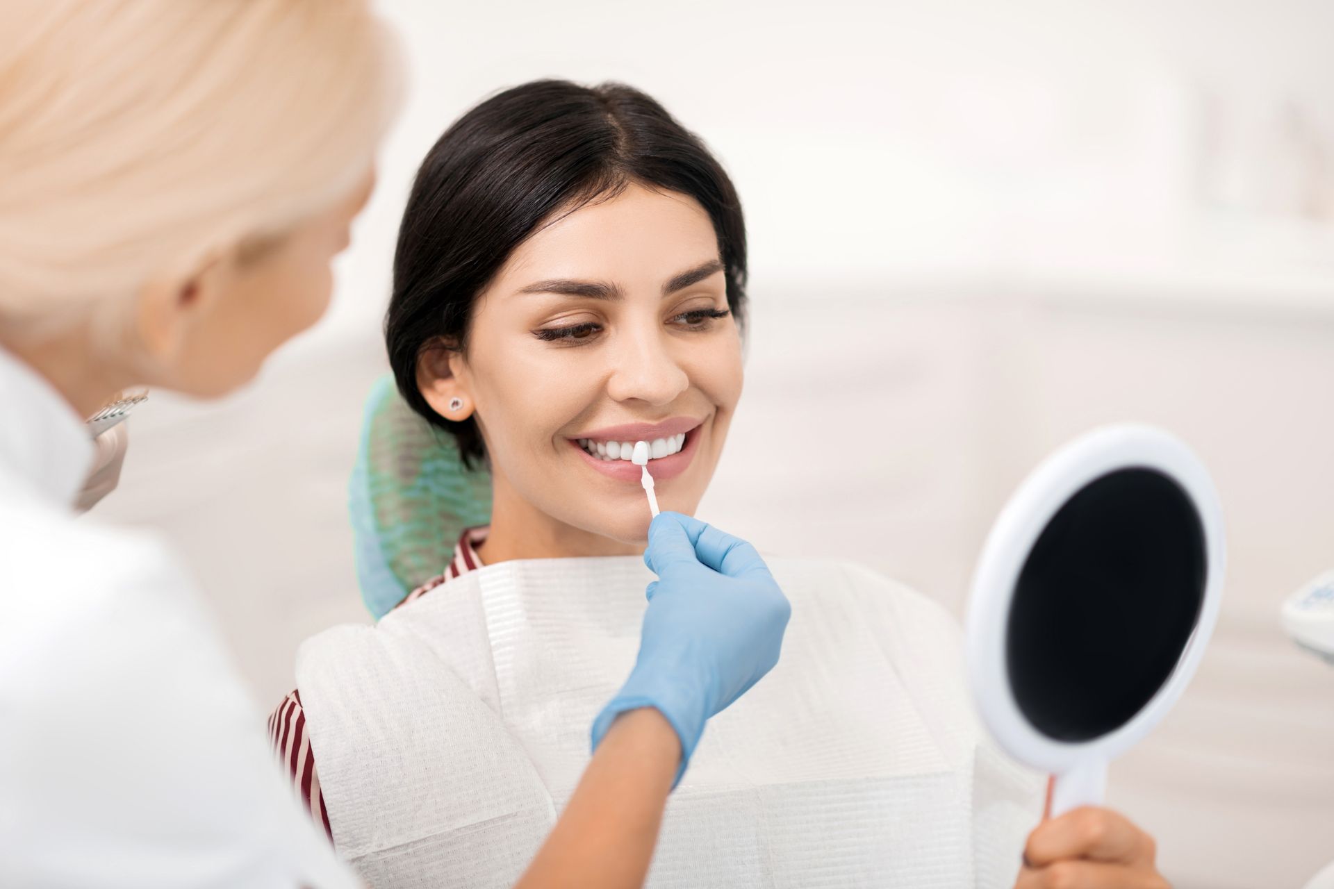 A woman is sitting in a dental chair looking at her teeth in a mirror.