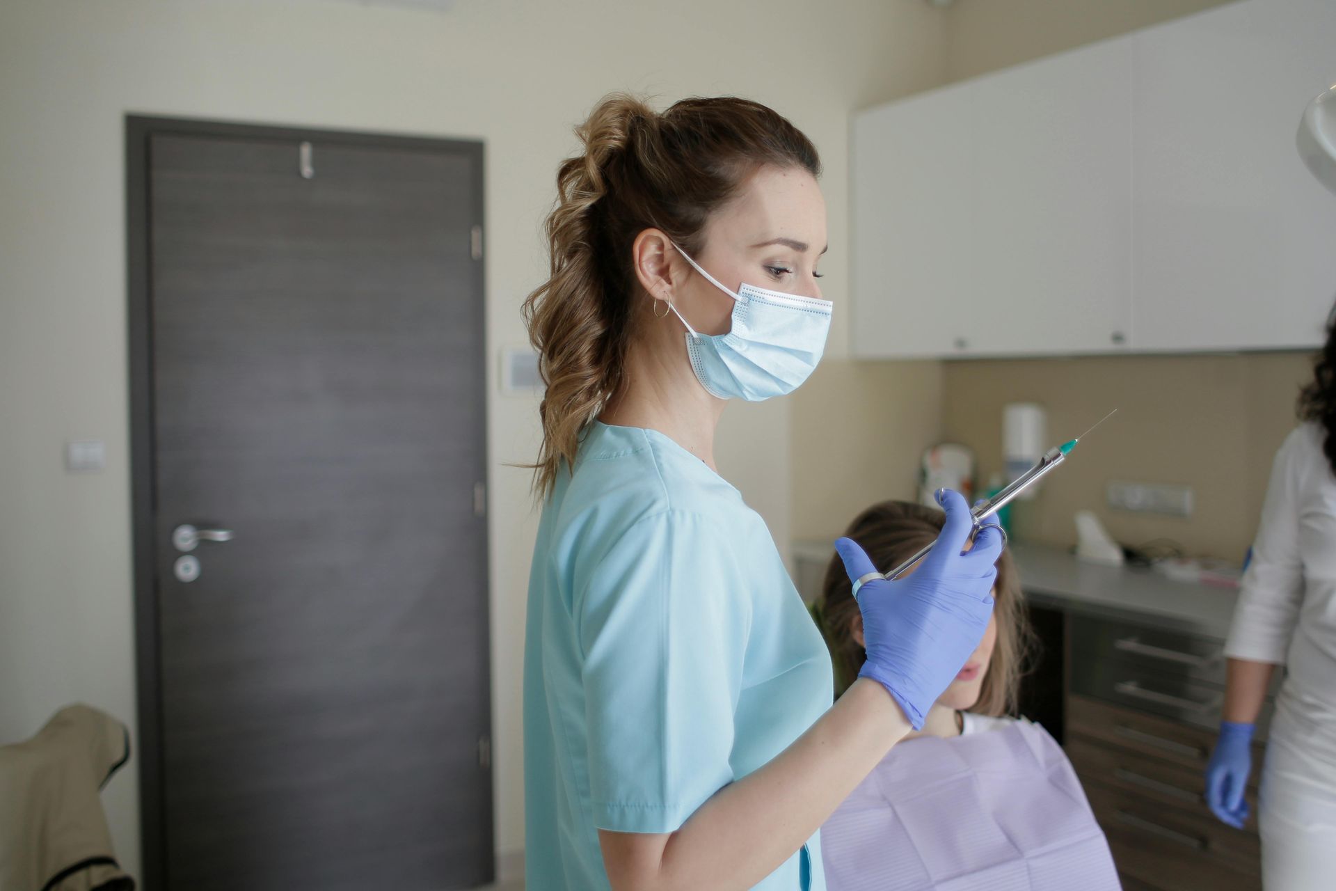 A female dentist wearing a mask and gloves is holding a syringe in front of a patient.