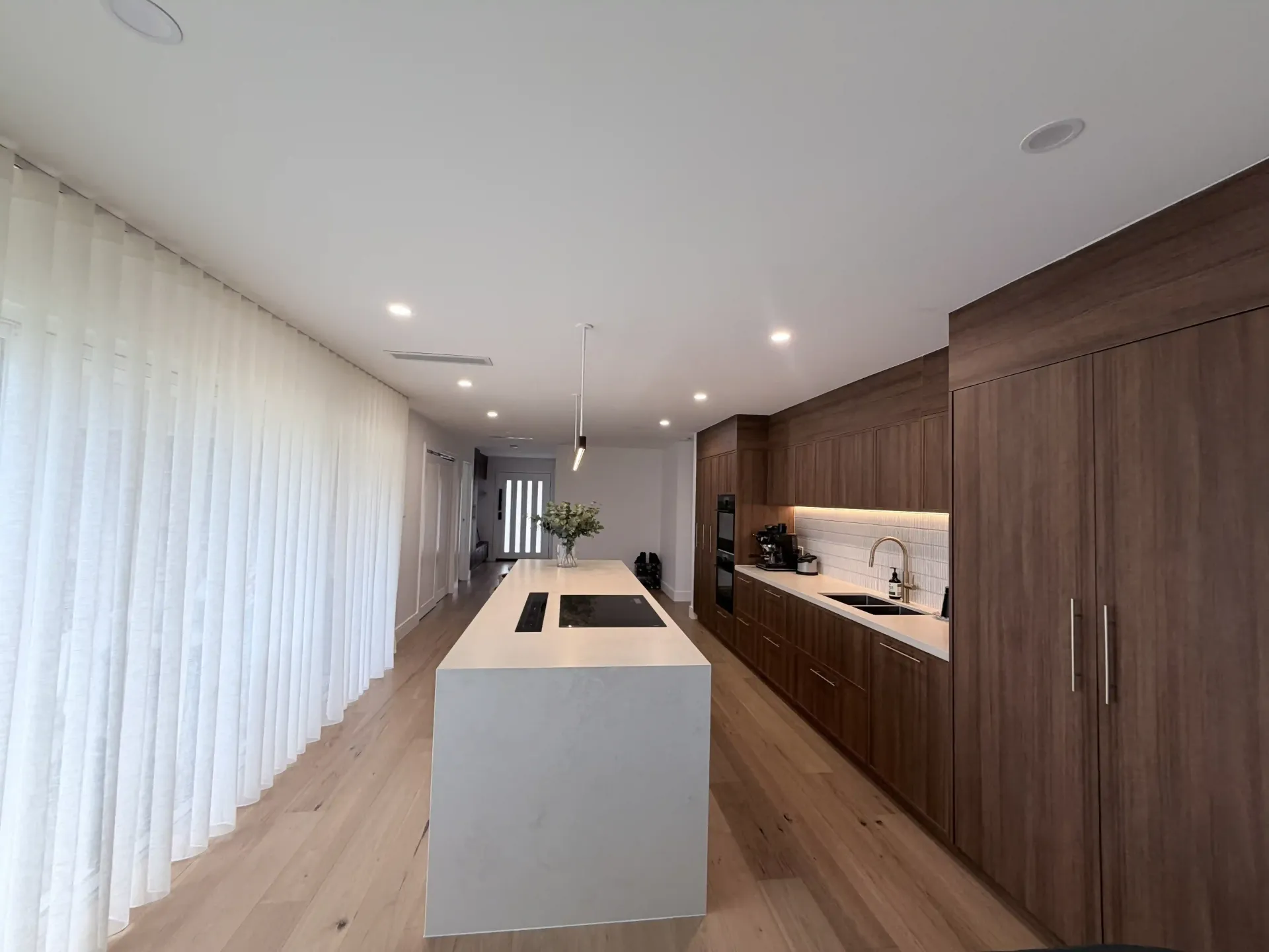Modern white kitchen with island, range hood, and windows; light wood floors.