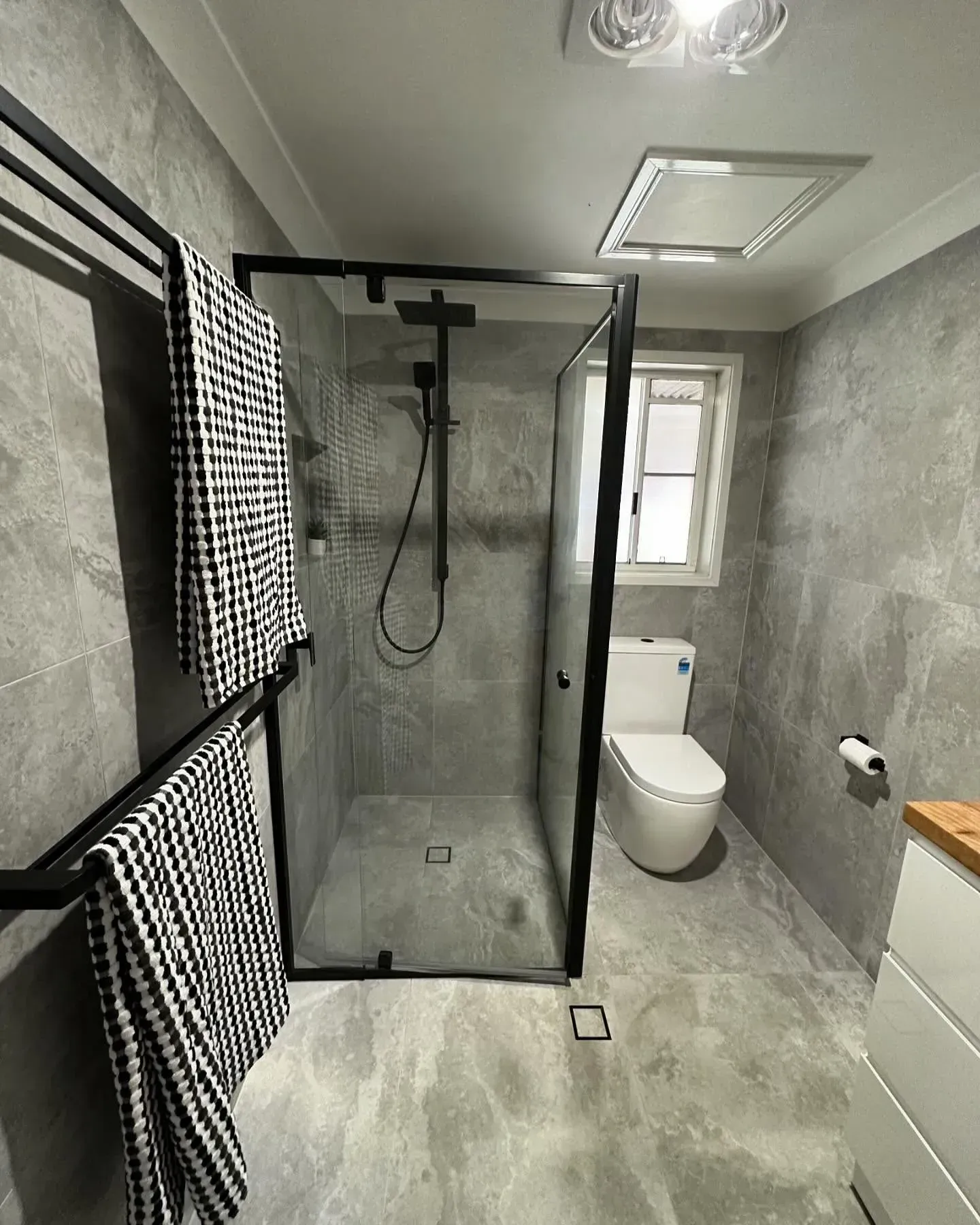 Modern bathroom with a glass shower, toilet, and gray tiled walls. Black towels hang on a rack.