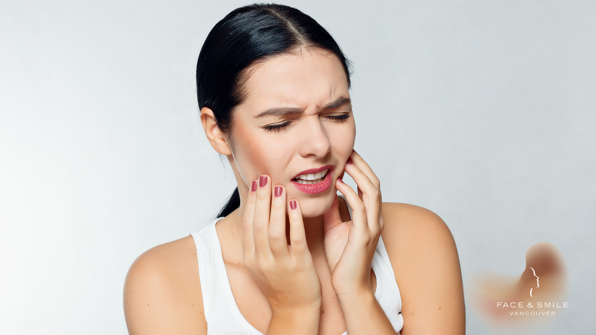 Woman holding face in pain, eyes closed, white tank top, light background.