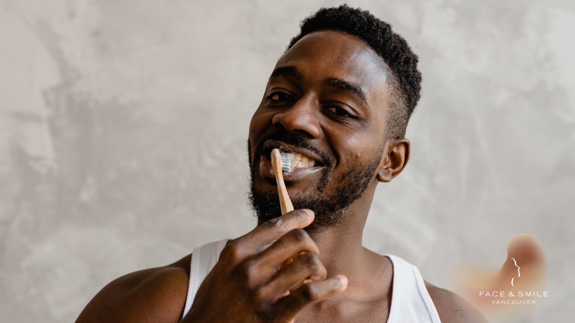 Man brushing teeth, smiling, in a white tank top, neutral background.
