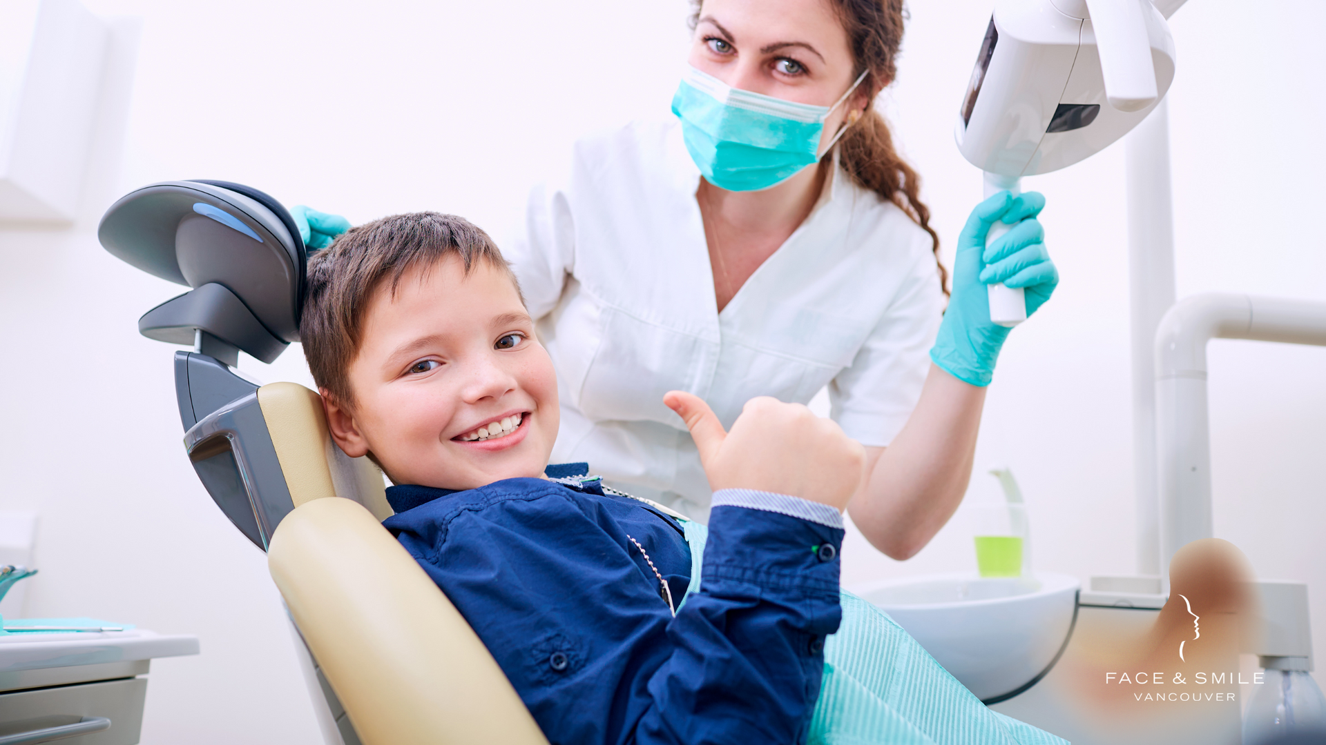 A young boy is sitting in a dental chair giving a thumbs up.