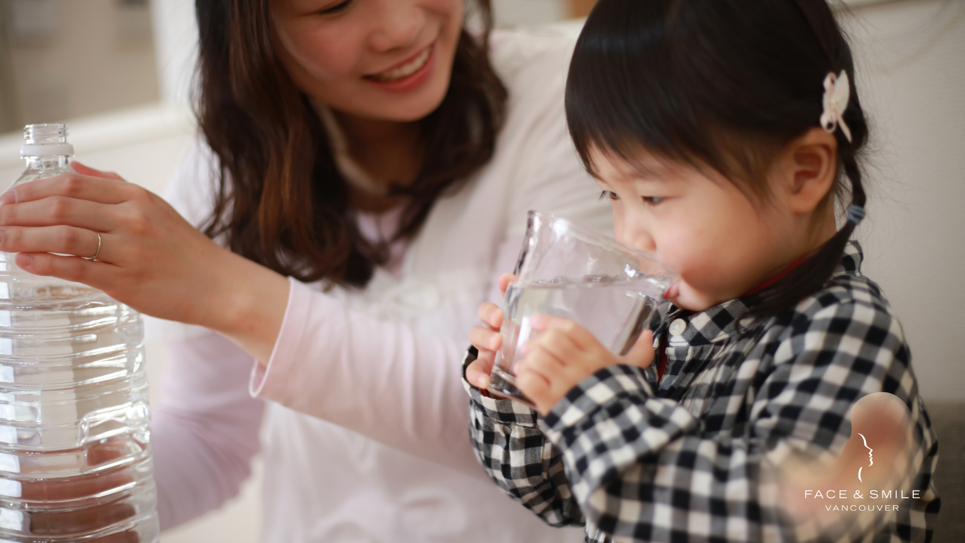 Woman pouring water for a child drinking from a glass. Indoors, natural light.