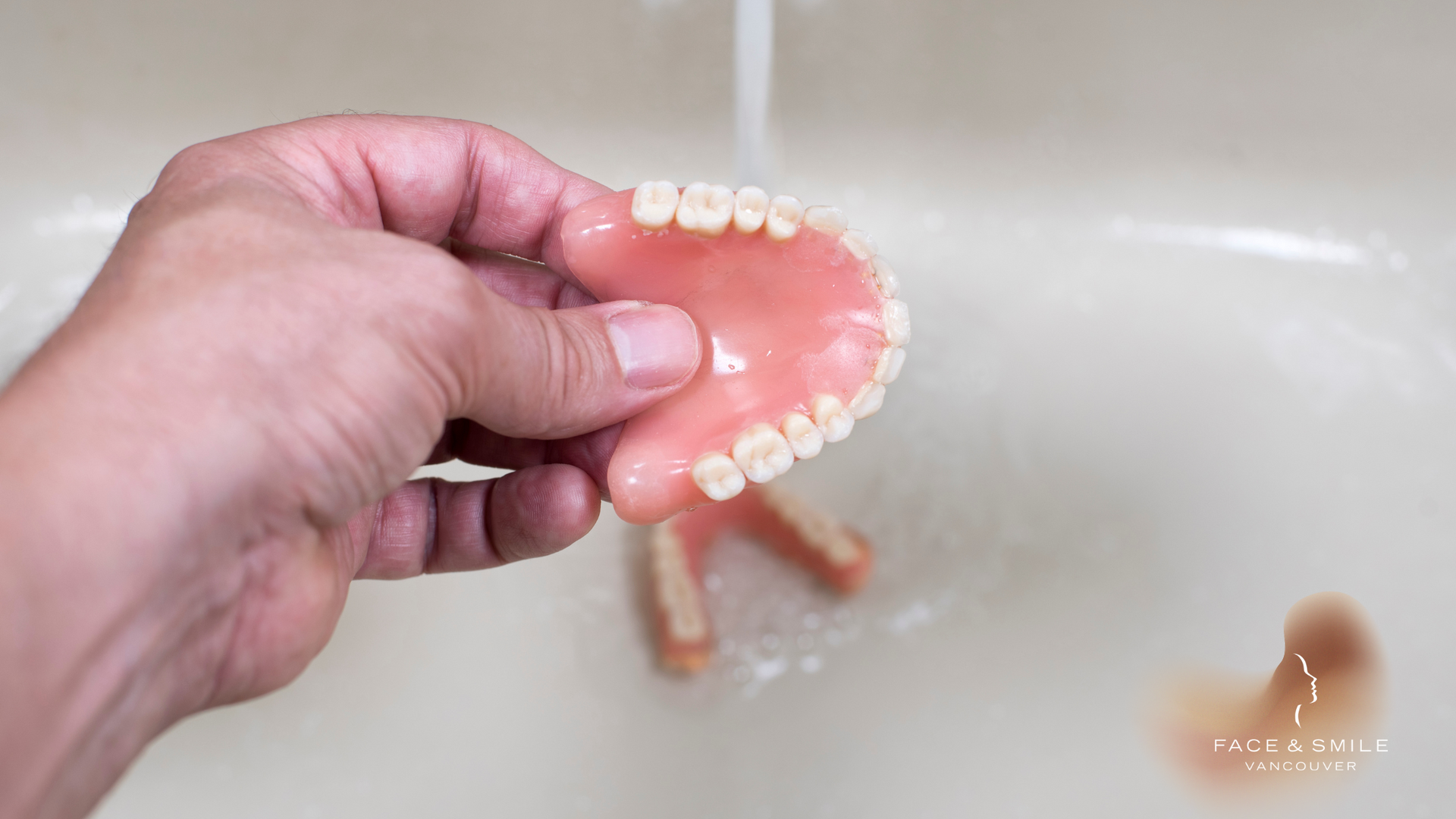 A person is holding a denture in their hand in front of a sink.