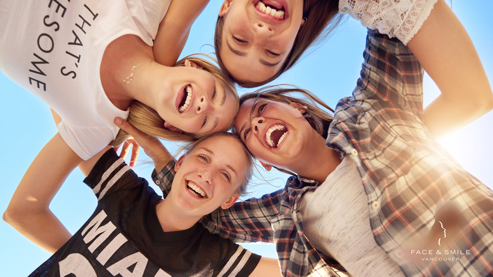 Four smiling people huddle together under a blue sky, arms around each other, looking down.