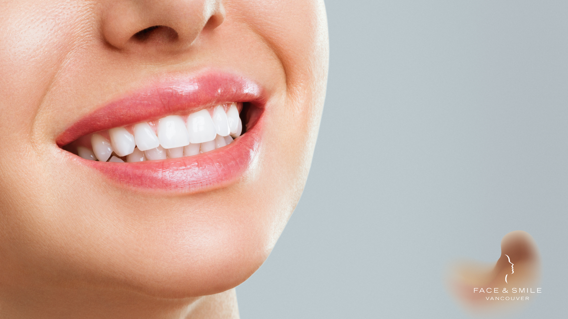 Close-up of a woman smiling, showcasing white teeth and pink lips against a gray background.