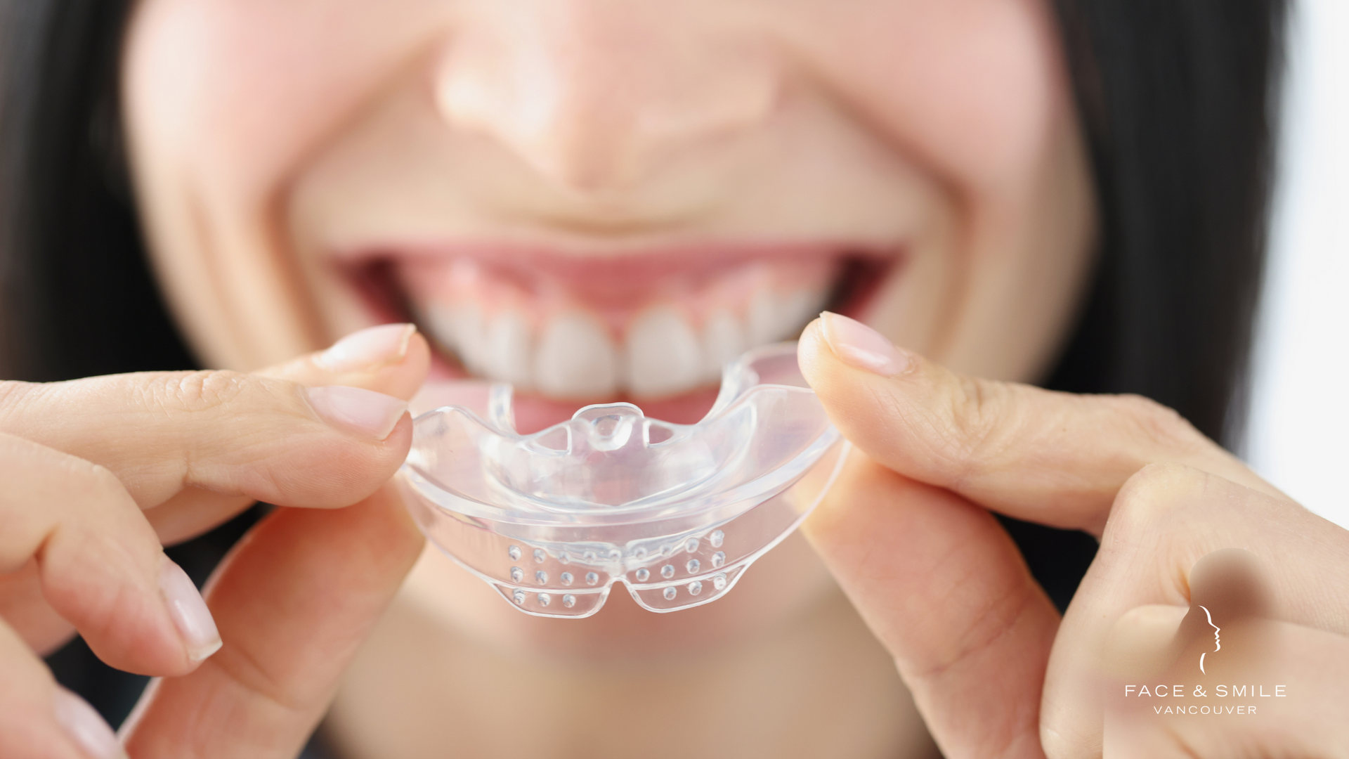 A woman is holding a clear retainer in front of her teeth.