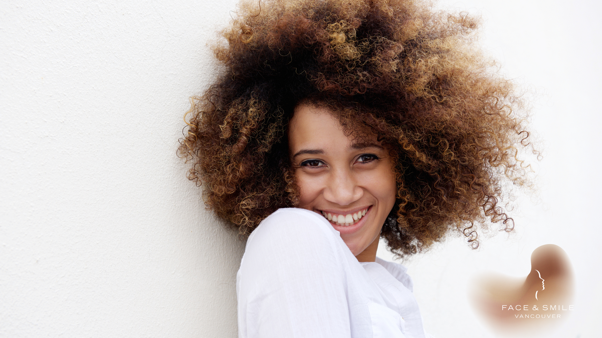 Smiling woman with large brown afro, leaning against a white wall.