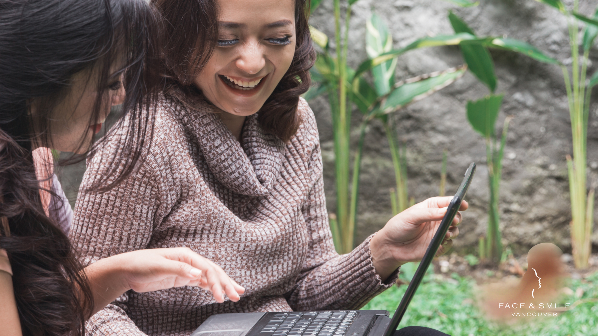 Two women are sitting on the grass looking at a laptop computer.