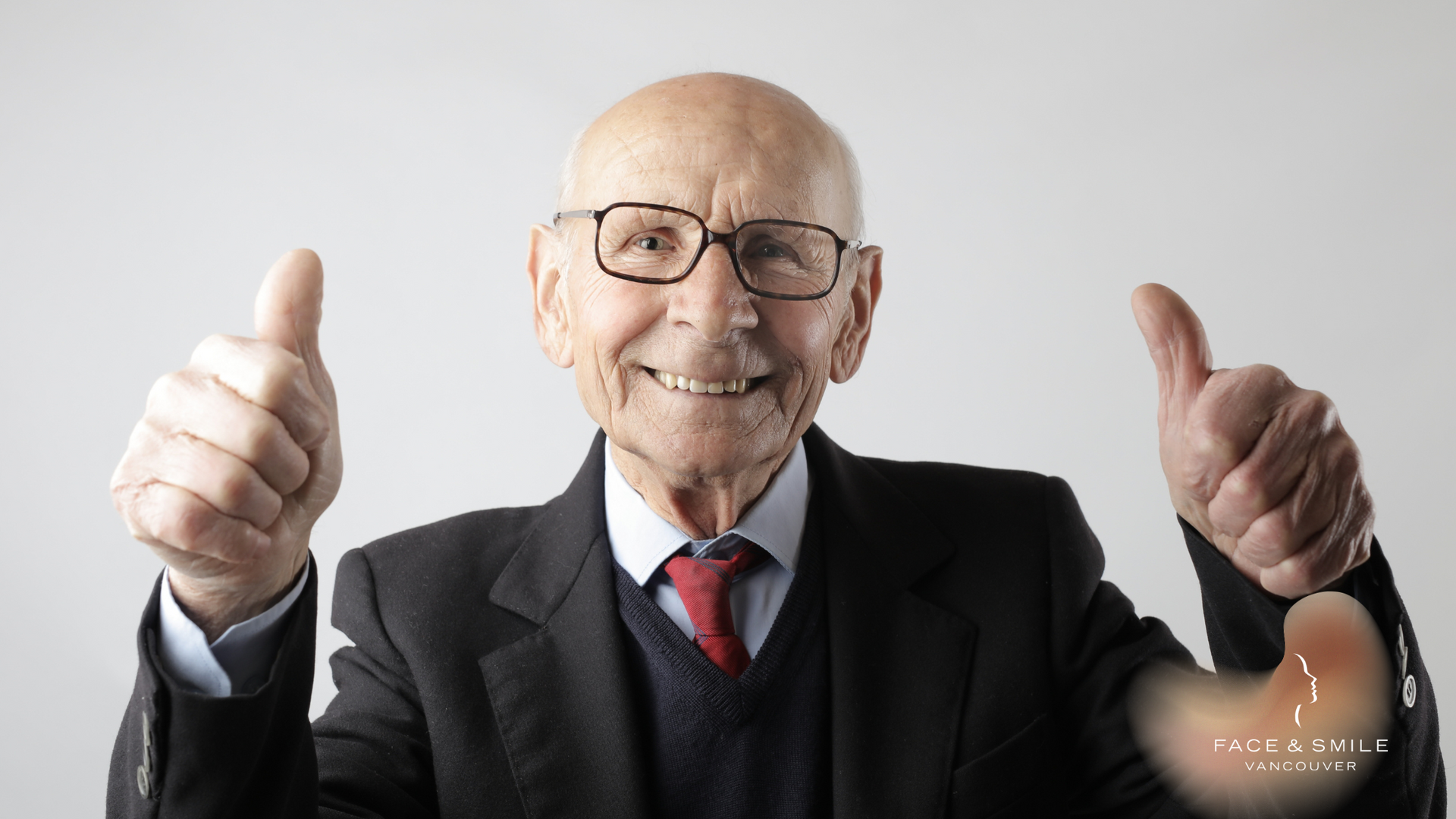 An elderly man in a suit and tie is giving two thumbs up.