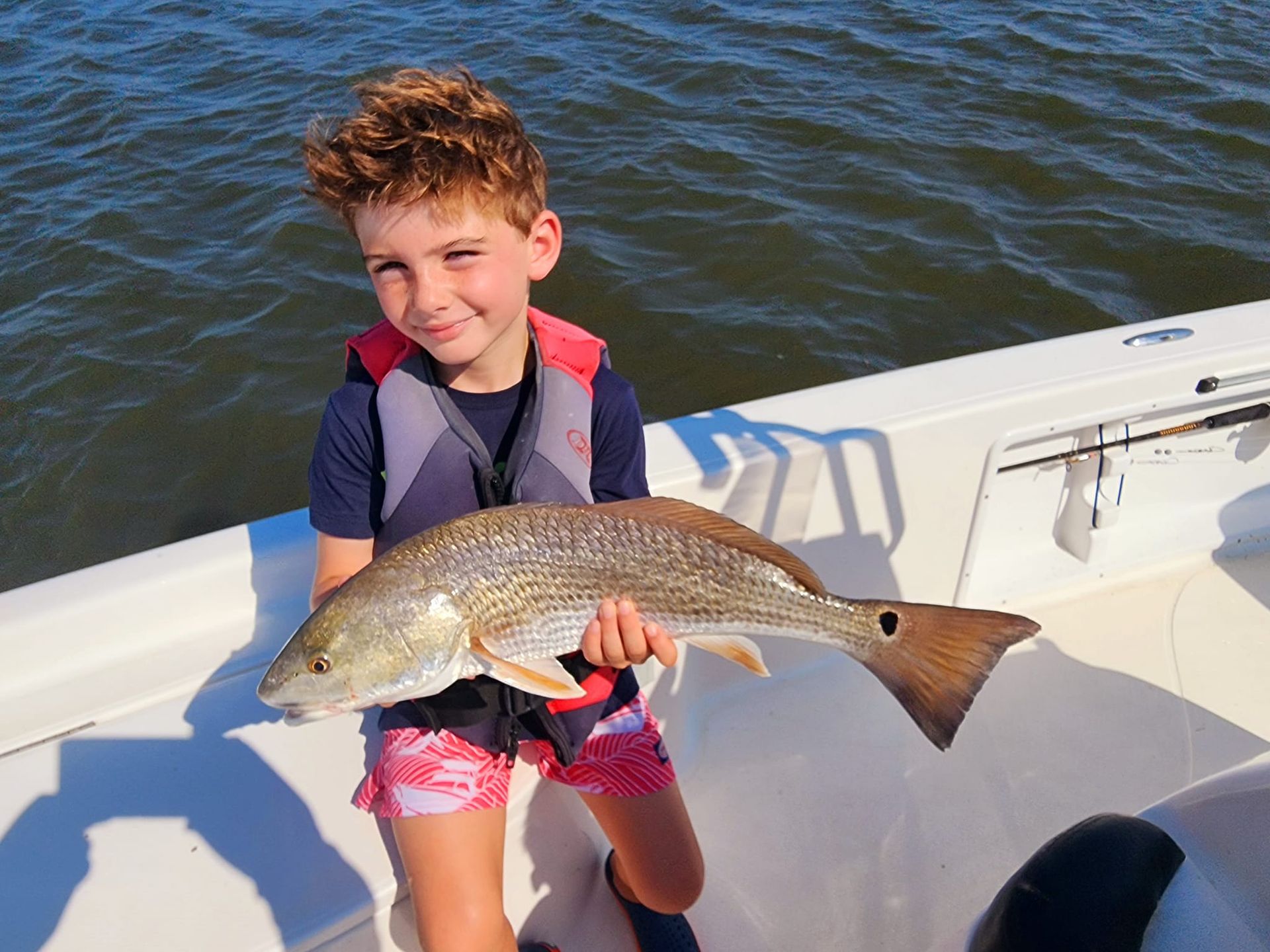 Boy on a boat smiles while holding a large redfish. Water and boat interior are visible.