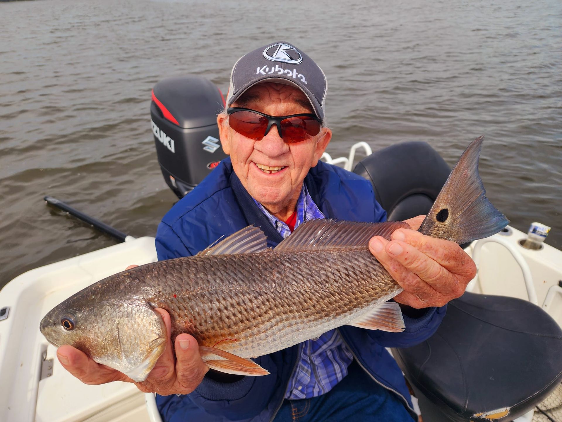 Man in sunglasses smiling, holding a redfish on a boat near an outboard motor.