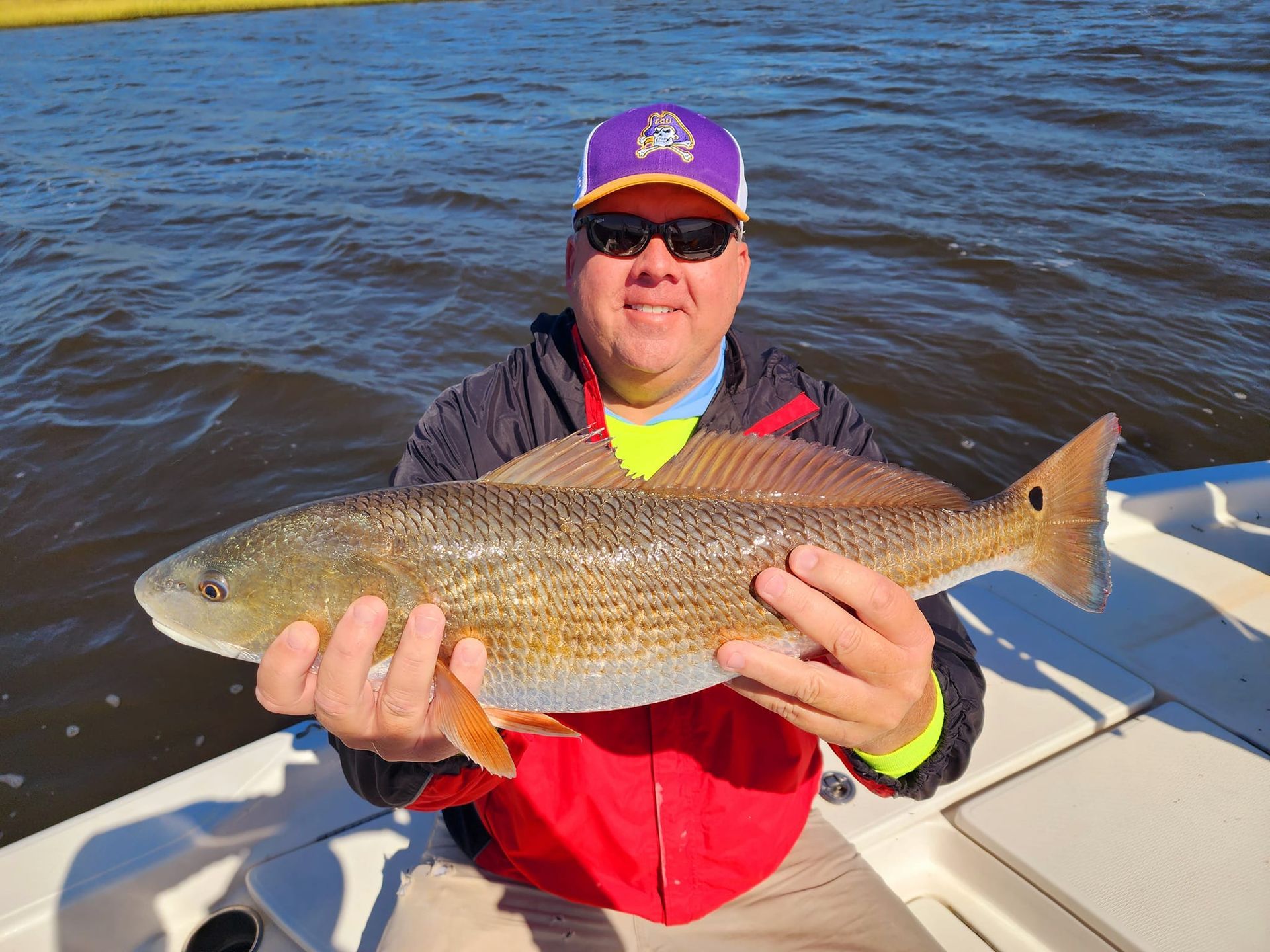 Man on a boat holds a large redfish. He's wearing sunglasses, a purple hat, and a red jacket, set against water.