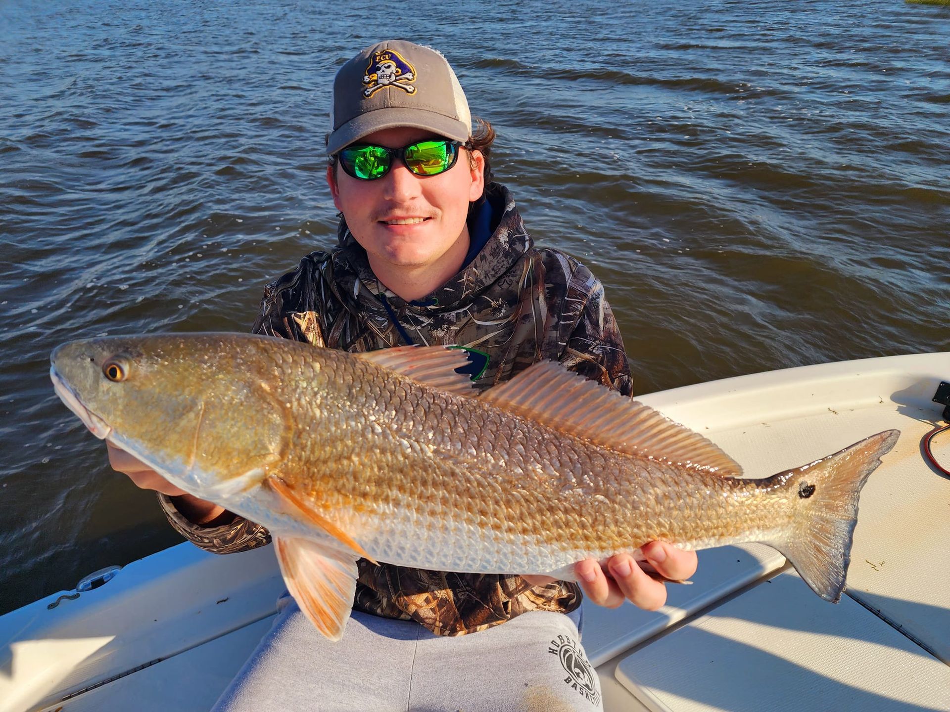 A person on a boat holds up a redfish. They are smiling, wearing sunglasses, a hat, and a camouflage jacket.