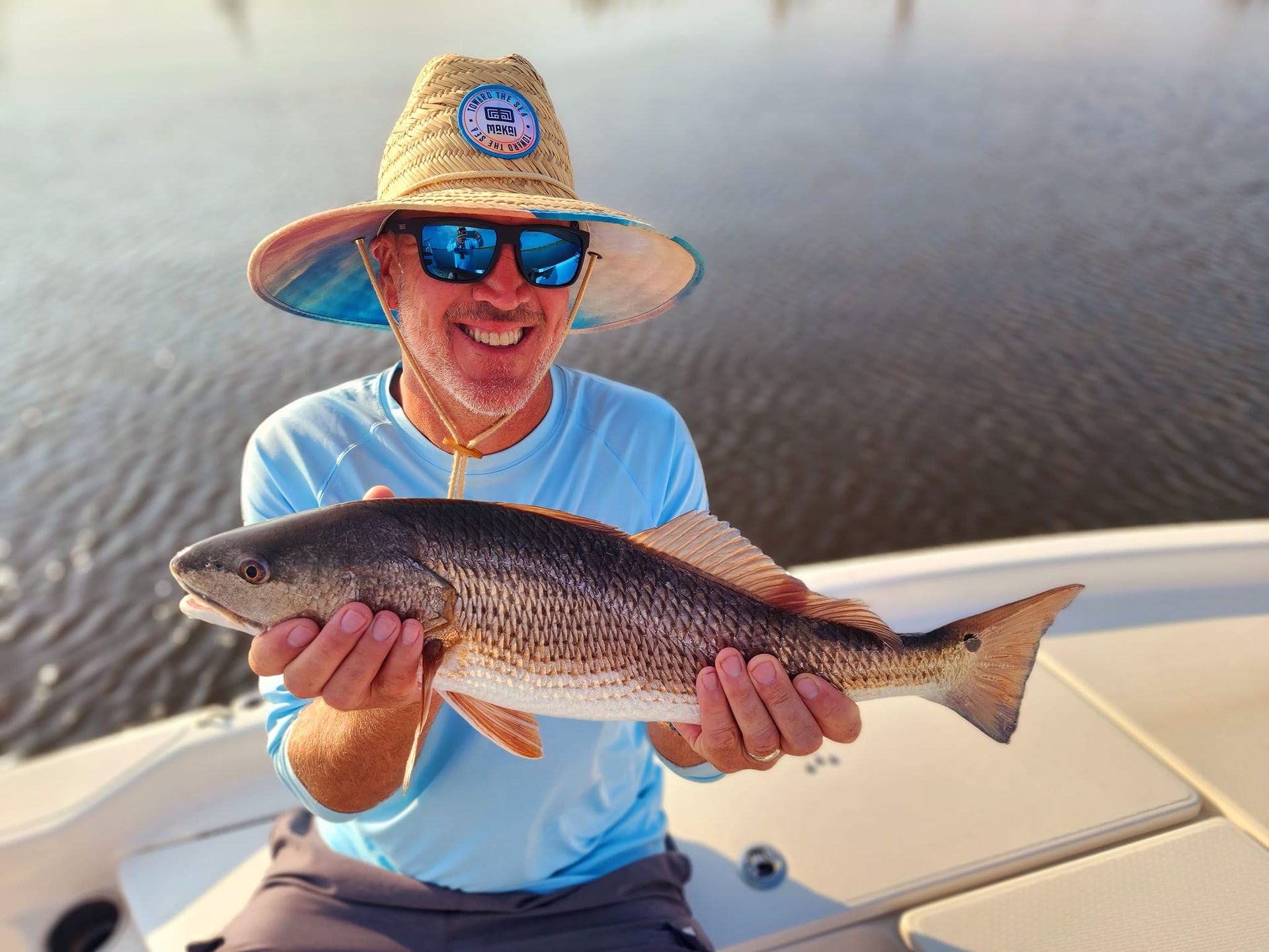 Man smiles, holding a redfish on a boat; water background. He wears a hat and sunglasses.