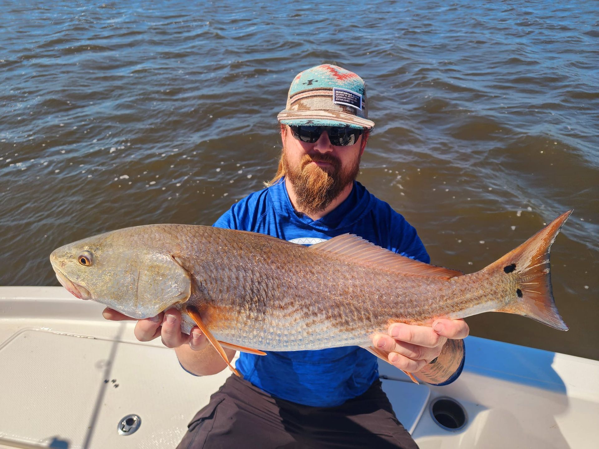 Man holding a large redfish on a boat, blue water and sky in background.