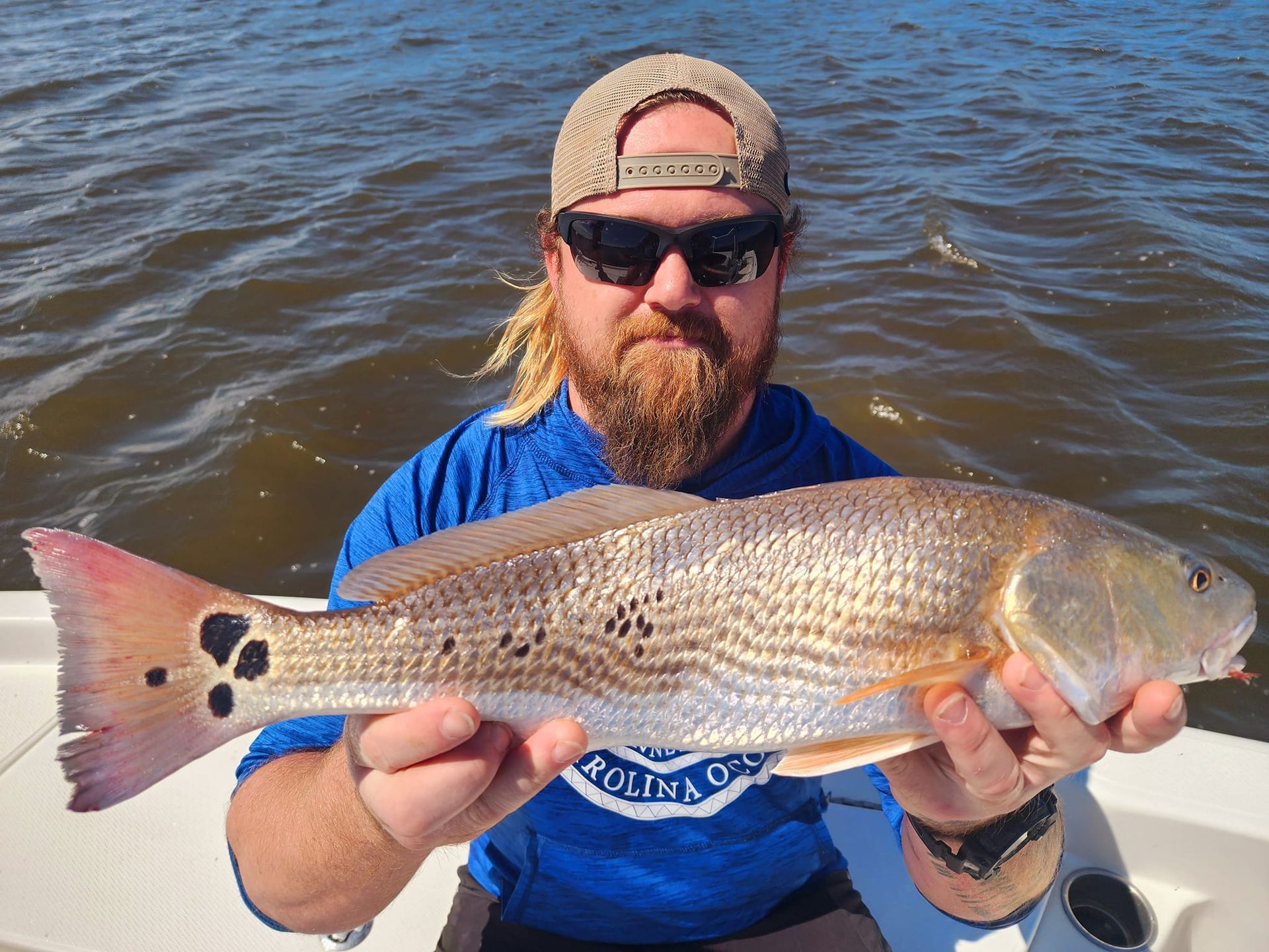 Man holding a redfish on a boat, wearing sunglasses and a cap, blue shirt.
