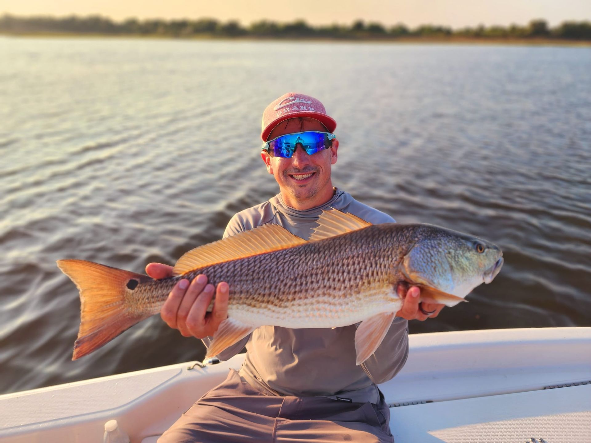 Man holding a large redfish on a boat, smiling, with water and shoreline visible.