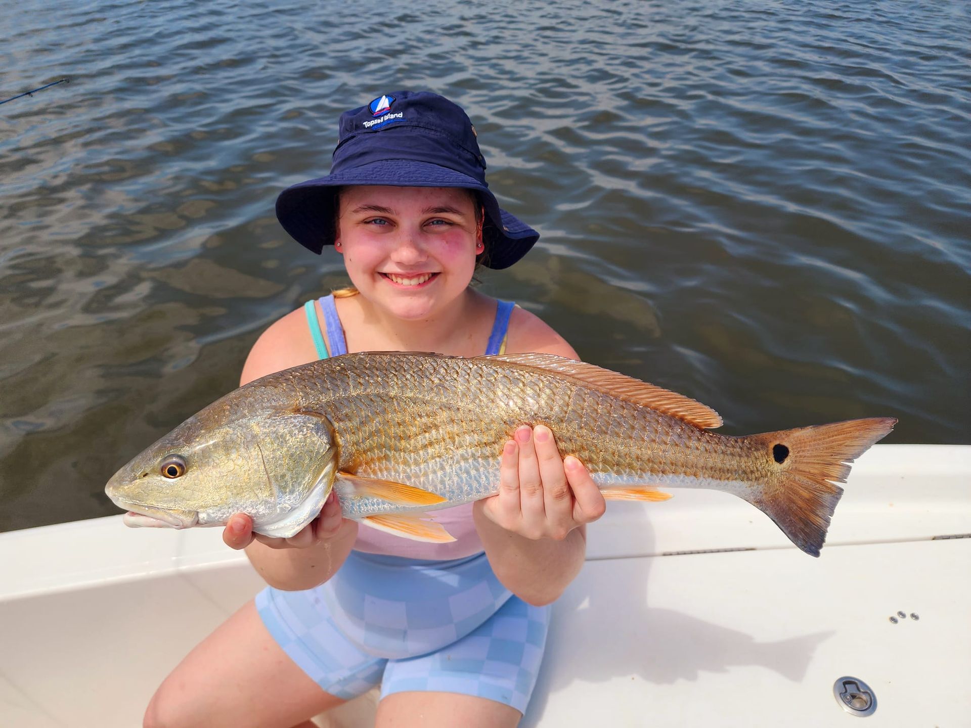 Young person holding a reddish-brown fish on a boat. The person is smiling and wearing a blue hat.