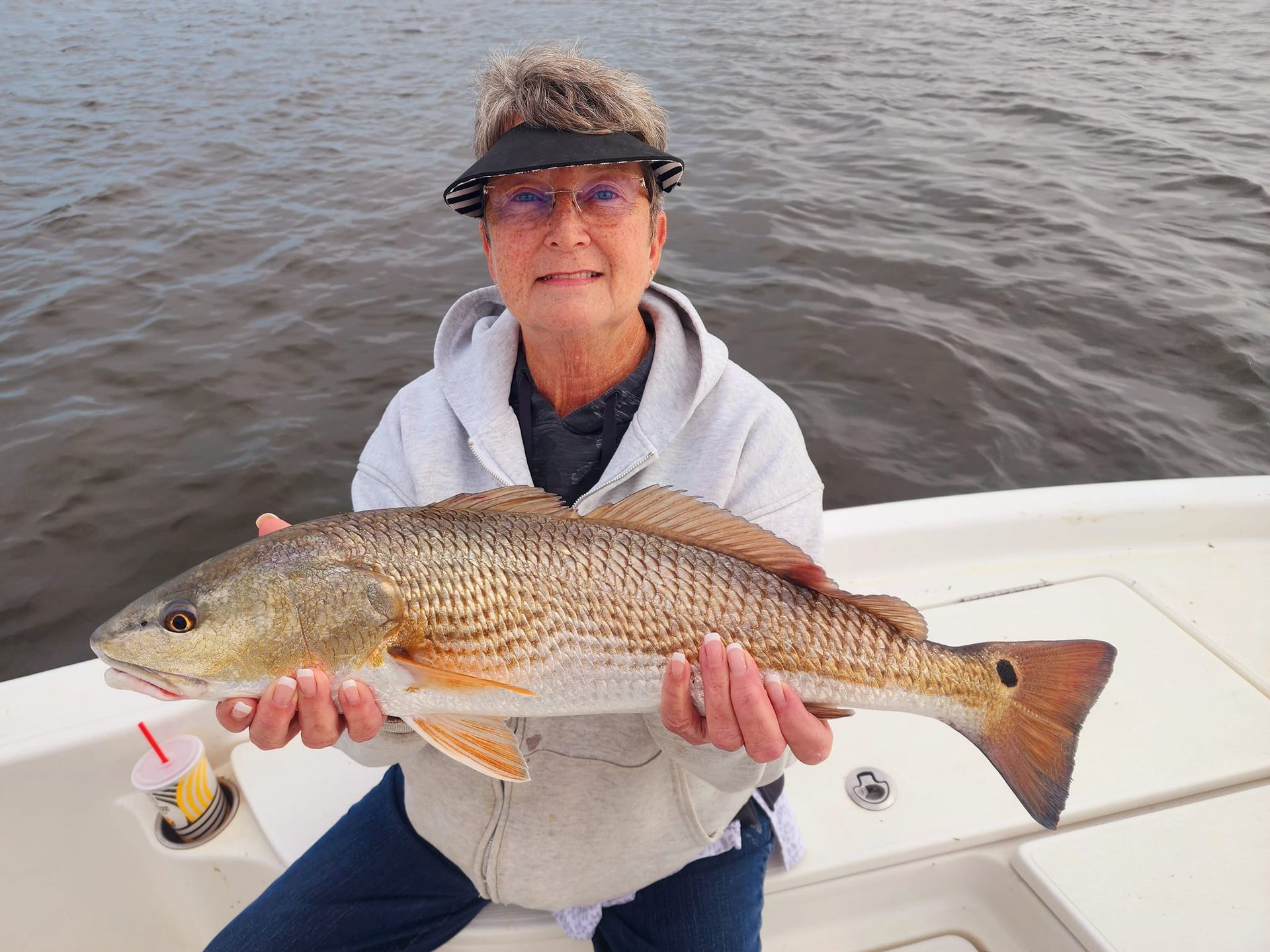 Woman on a boat holding a large redfish. Gray water, sunny visor, white sweatshirt.