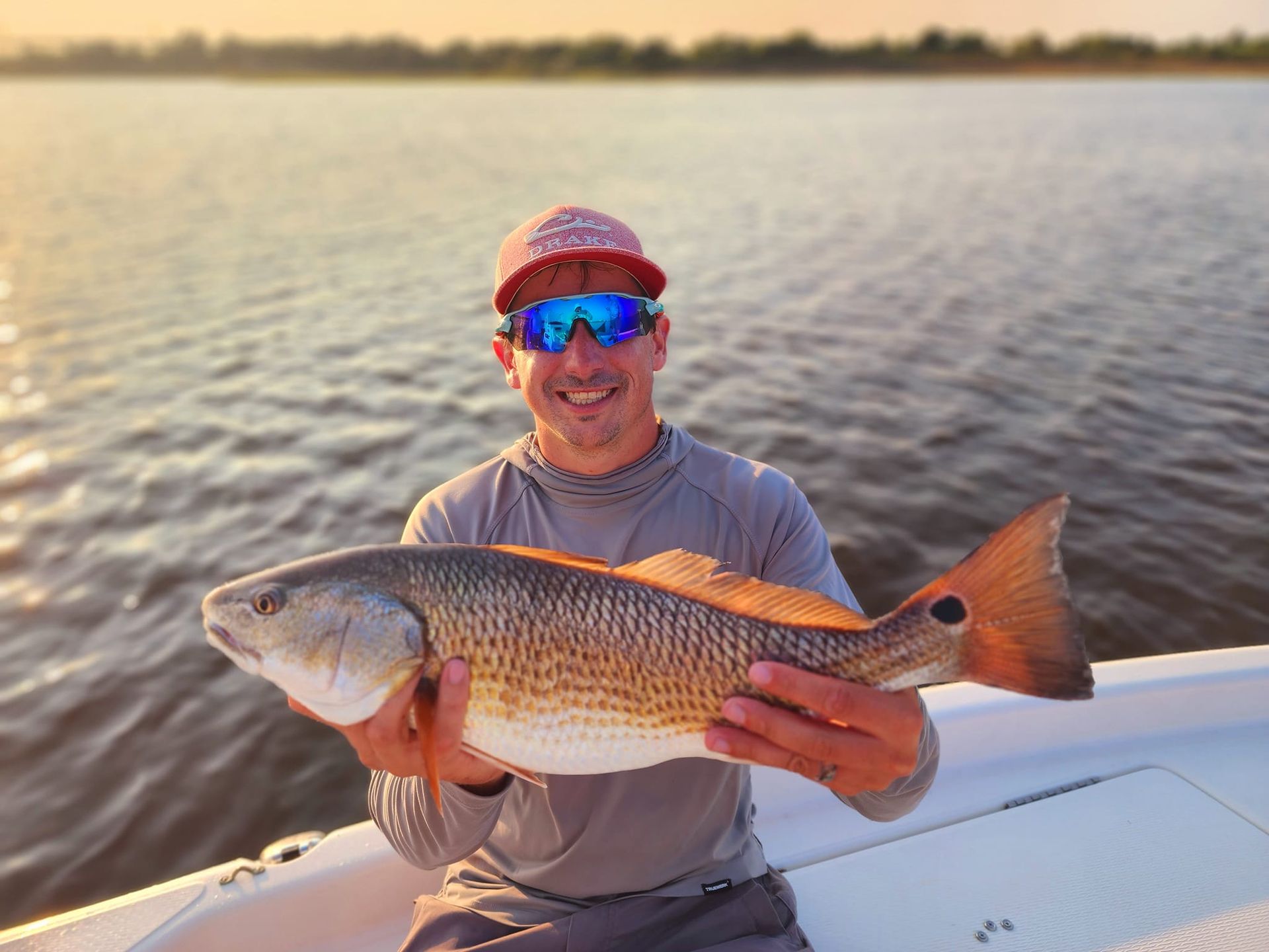 Man on a boat holding a redfish, sunset over water in the background.