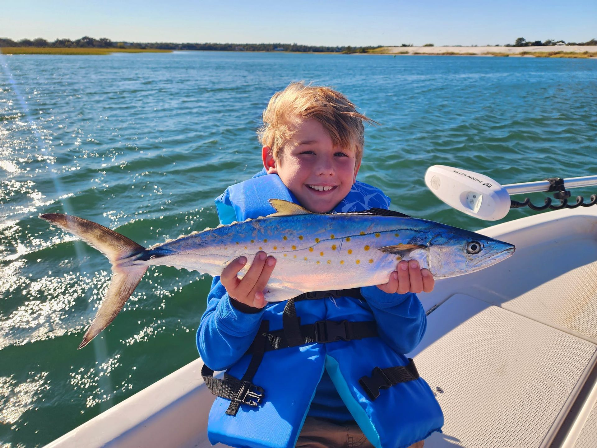 Boy in blue life vest holding a fish on a boat, smiling, with ocean background.
