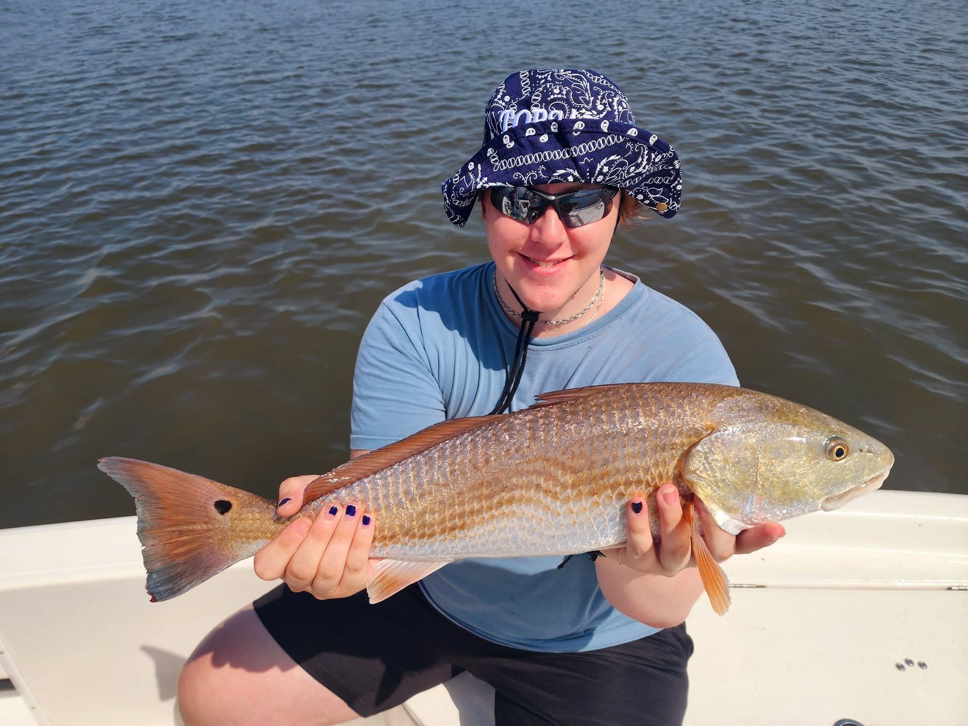 Person holding a large reddish-bronze fish on a boat. The person wears a blue hat and sunglasses, smiling.