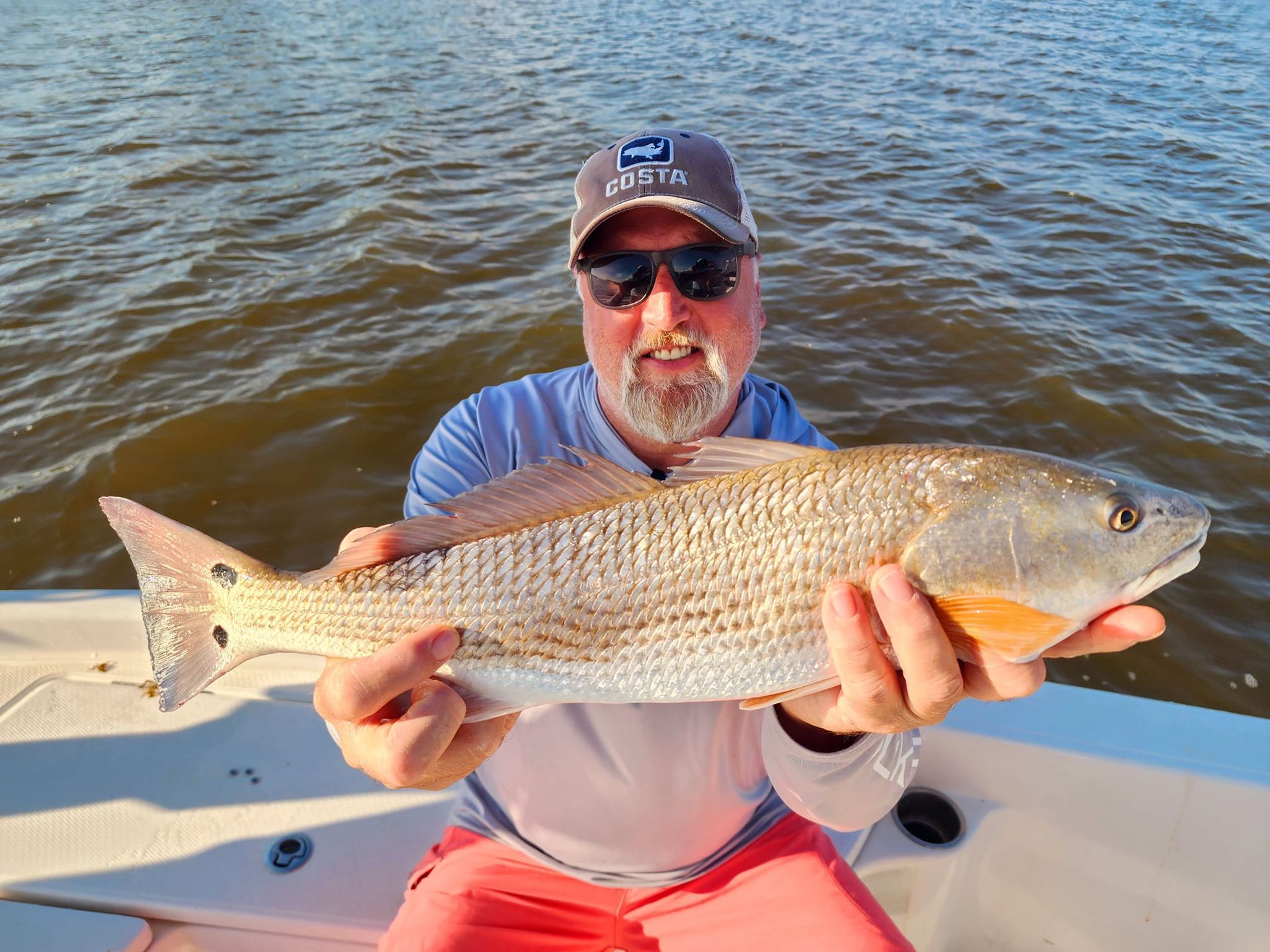 Man in sunglasses holding a redfish on a boat; water in the background.
