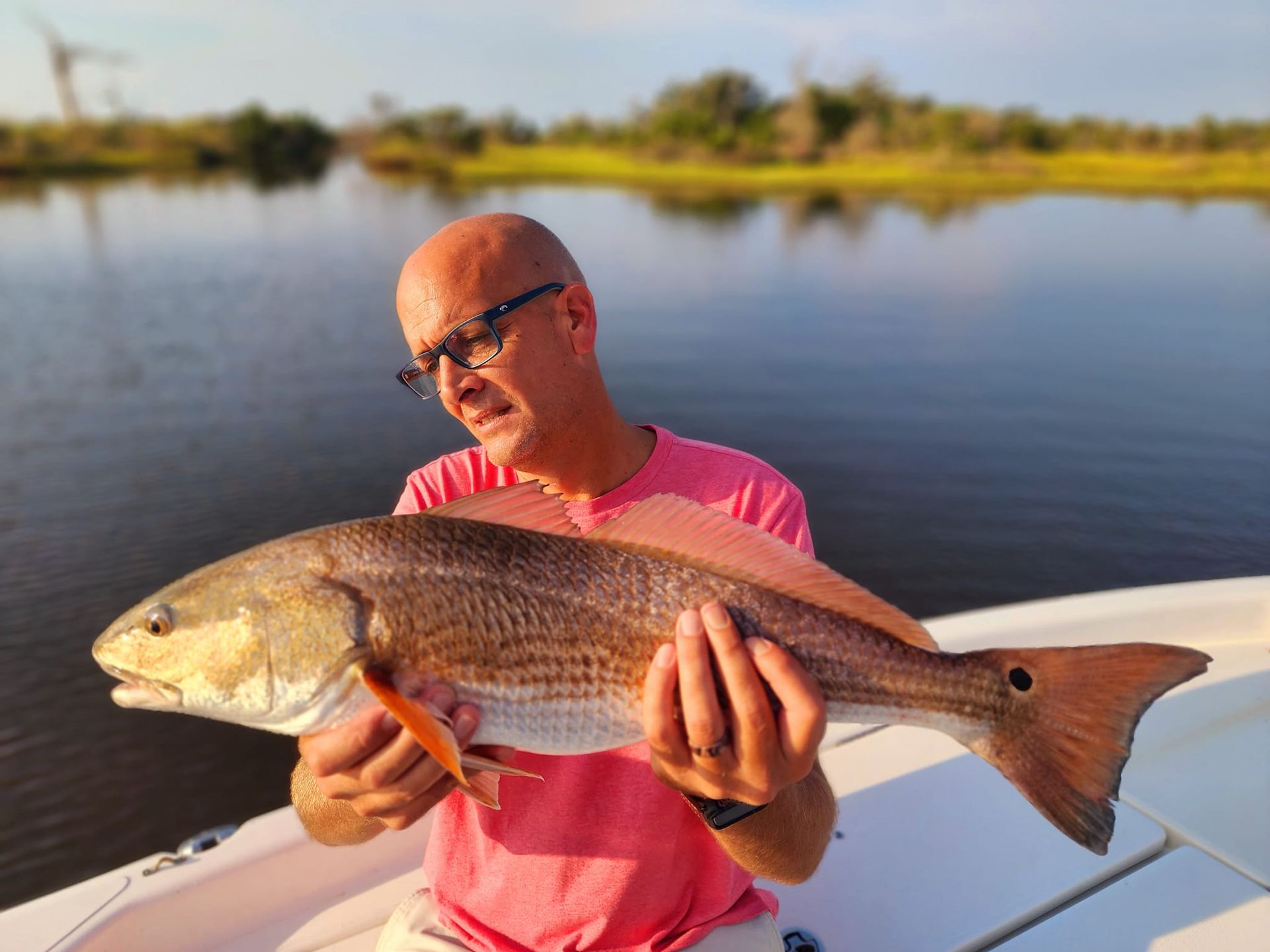 Man holding large redfish on a boat, near a calm waterway, sunny day.