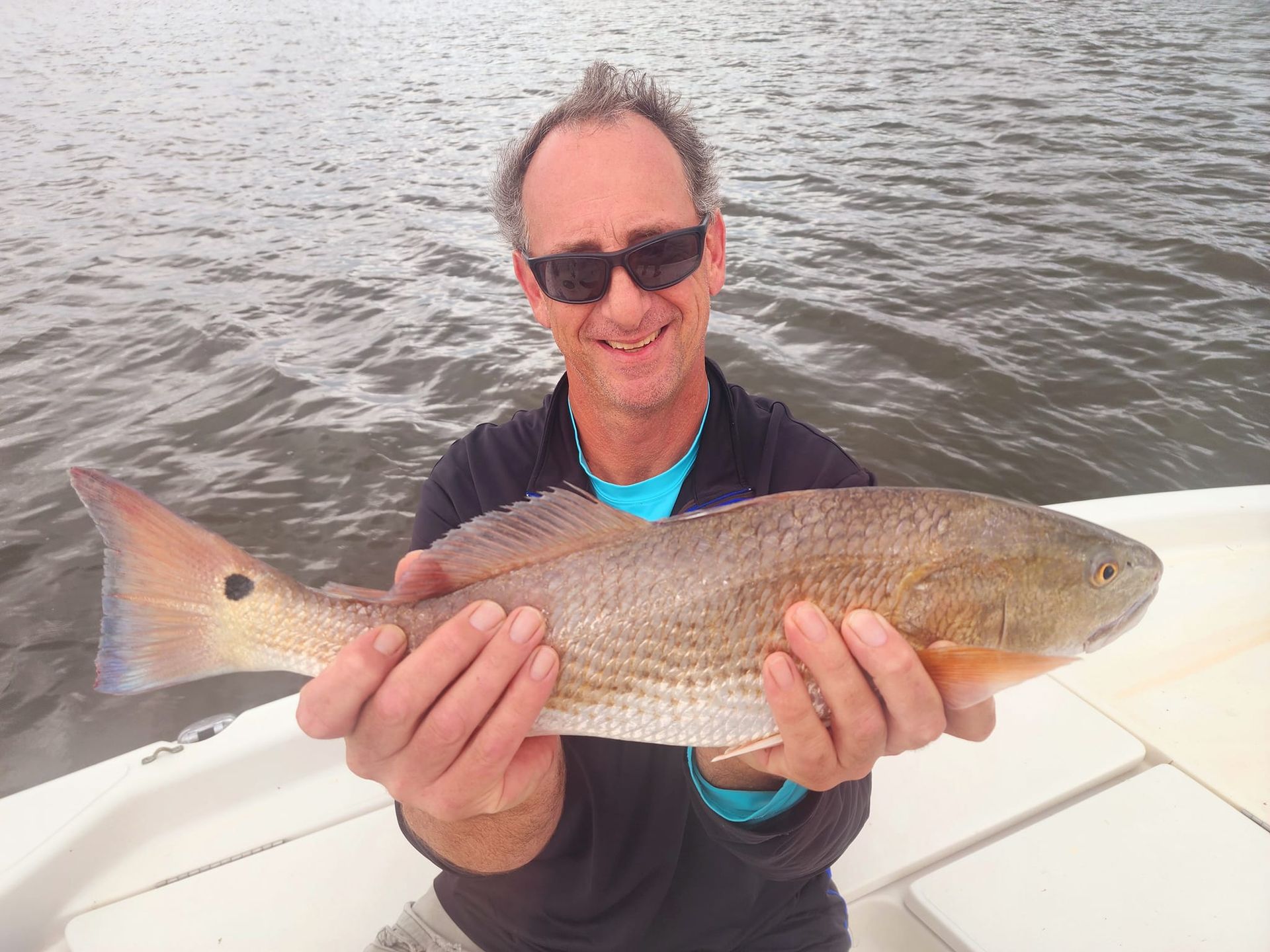 Man holding a reddish fish on a boat. The man is smiling. Cloudy day.